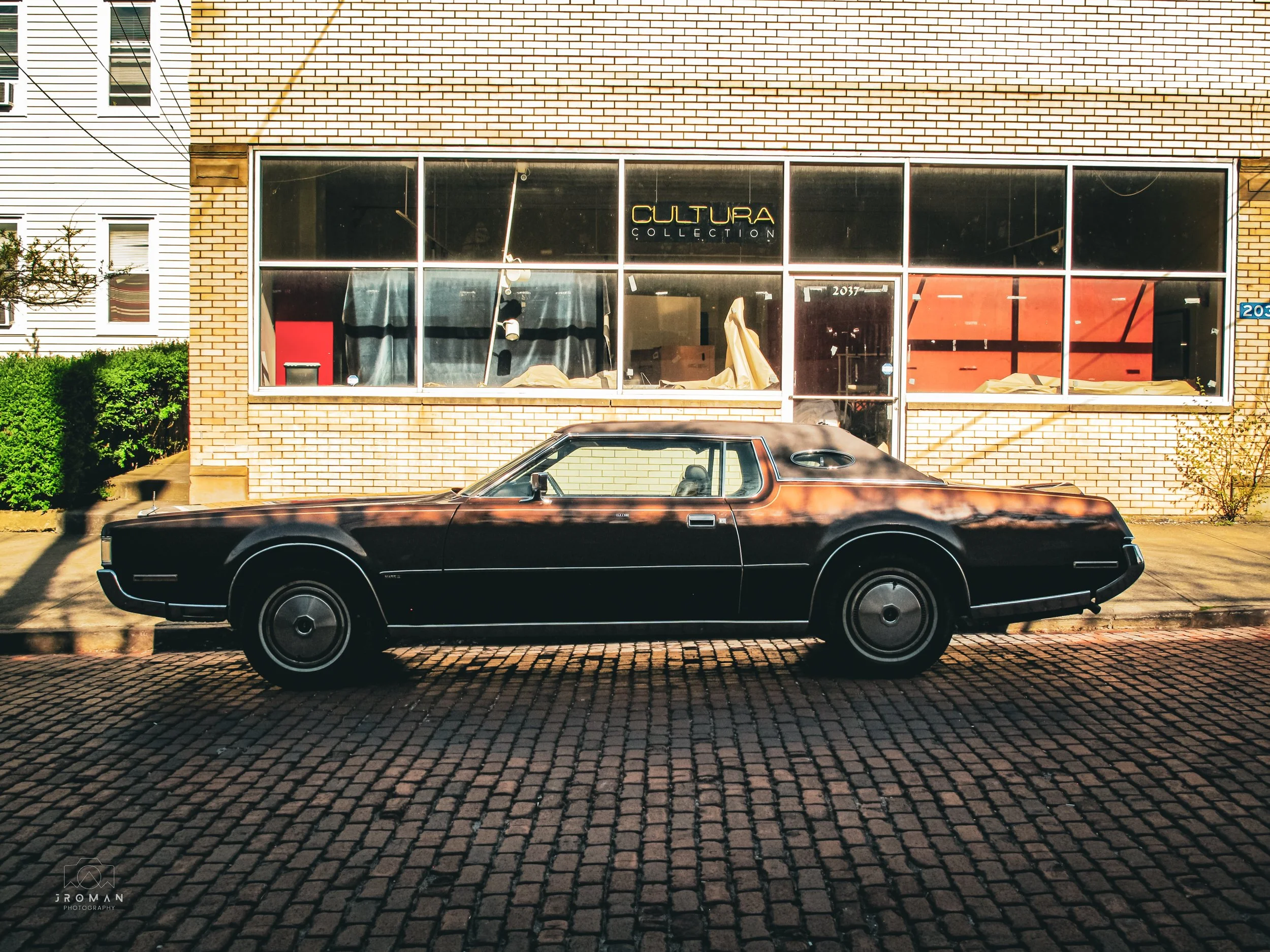 A black vintage car parked on cobblestone street in front of a brick building with large windows displaying a sign that reads "CULTURA COLLECTION."