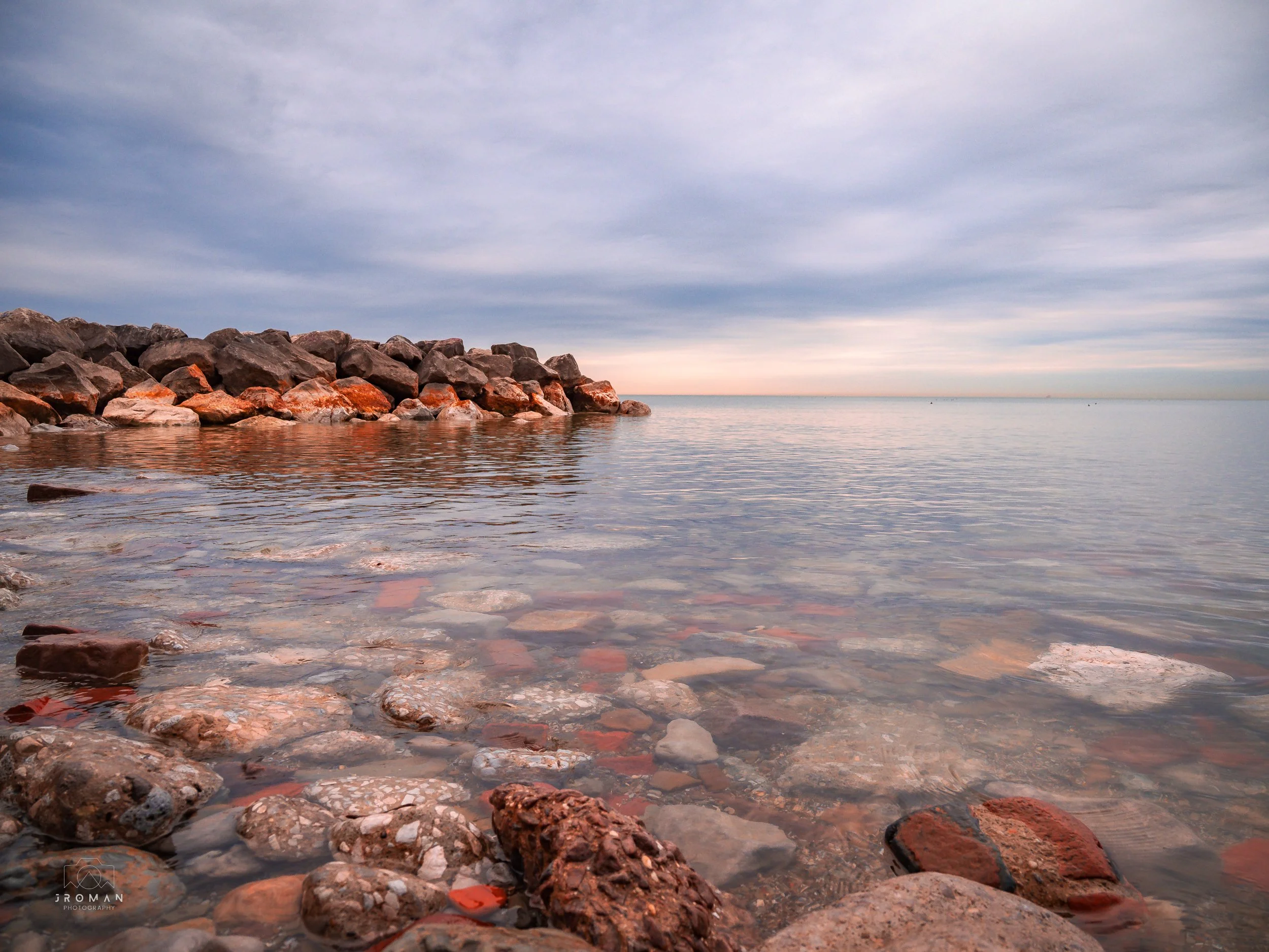 Calm ocean waters with a rocky shoreline in the foreground and a breakwater made of large rocks on the left. The sky is partly cloudy with soft, pastel-colored clouds and a faint horizon line in the distance.
