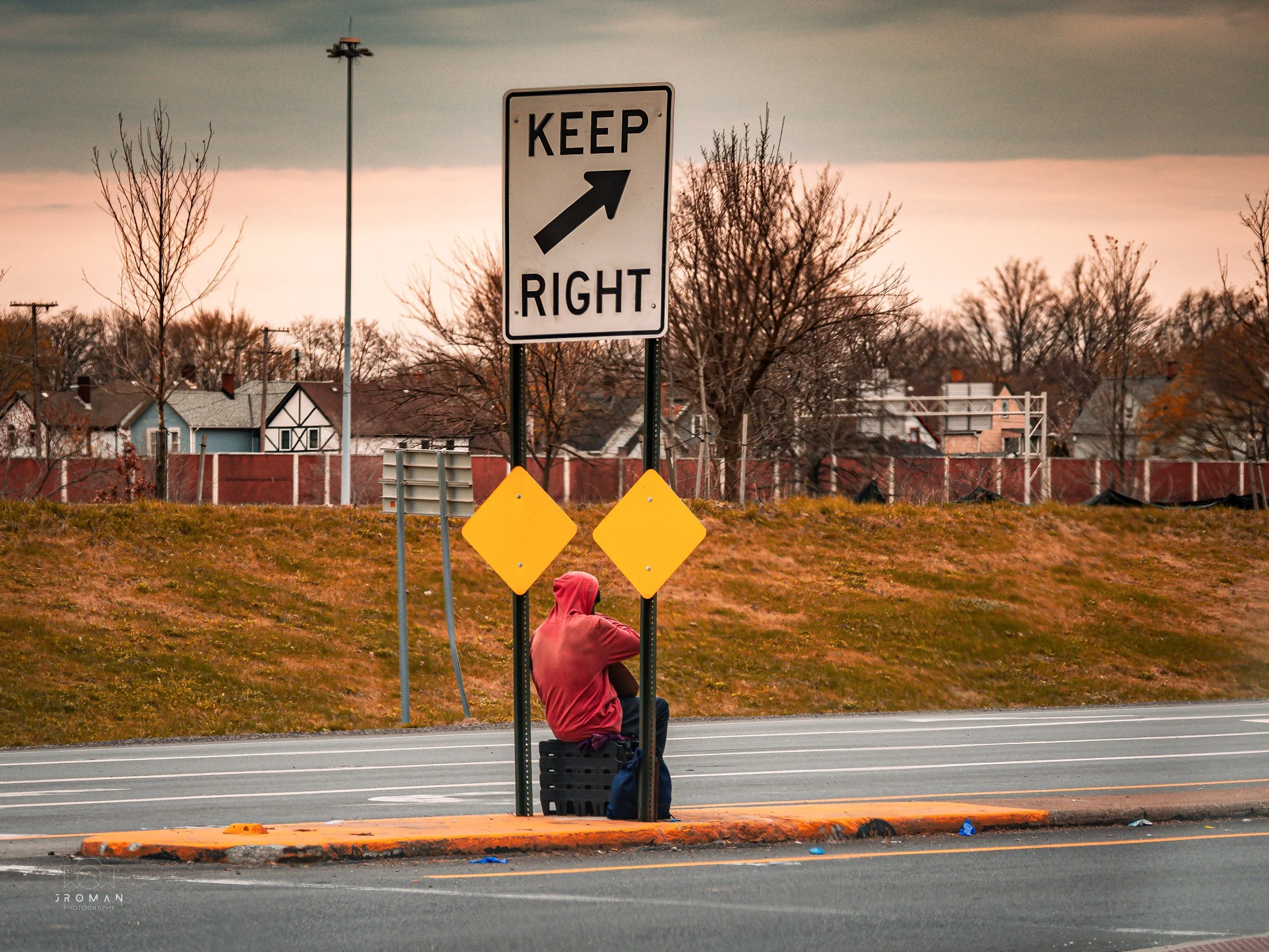 A person sitting at a bus stop on the side of the road, wearing a pink hoodie, with a sign that says 'Keep Right' behind them. The background shows houses, trees, and a cloudy sky.