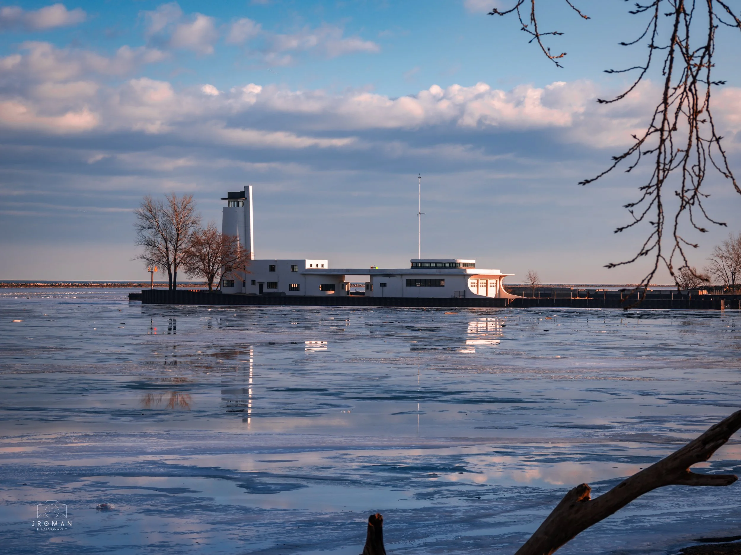 A building located by a frozen body of water, with leafless trees on either side and a cloudy sky above.
