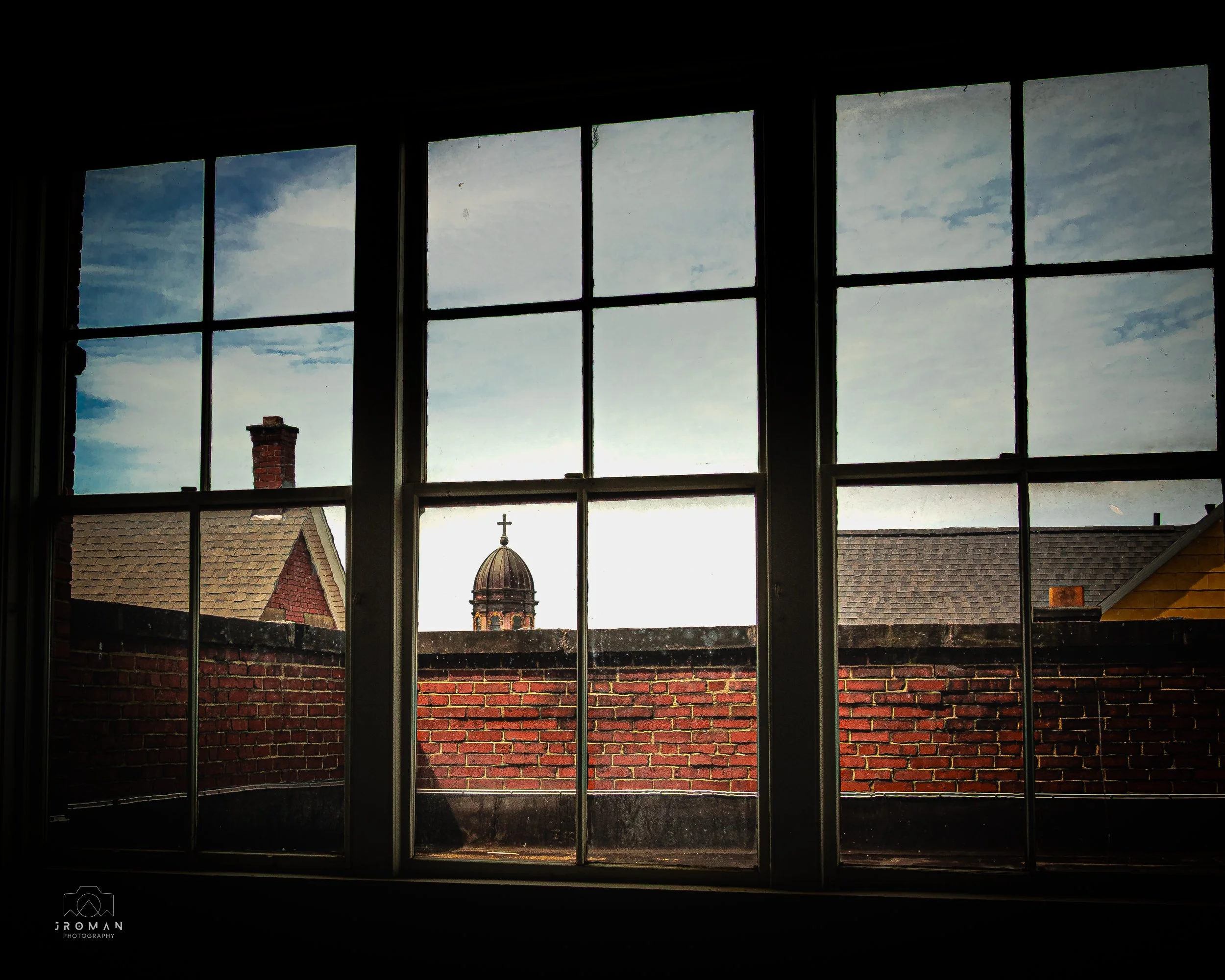 View through a window showing a brick rooftop, a church dome with a cross, and a cloudy sky.
