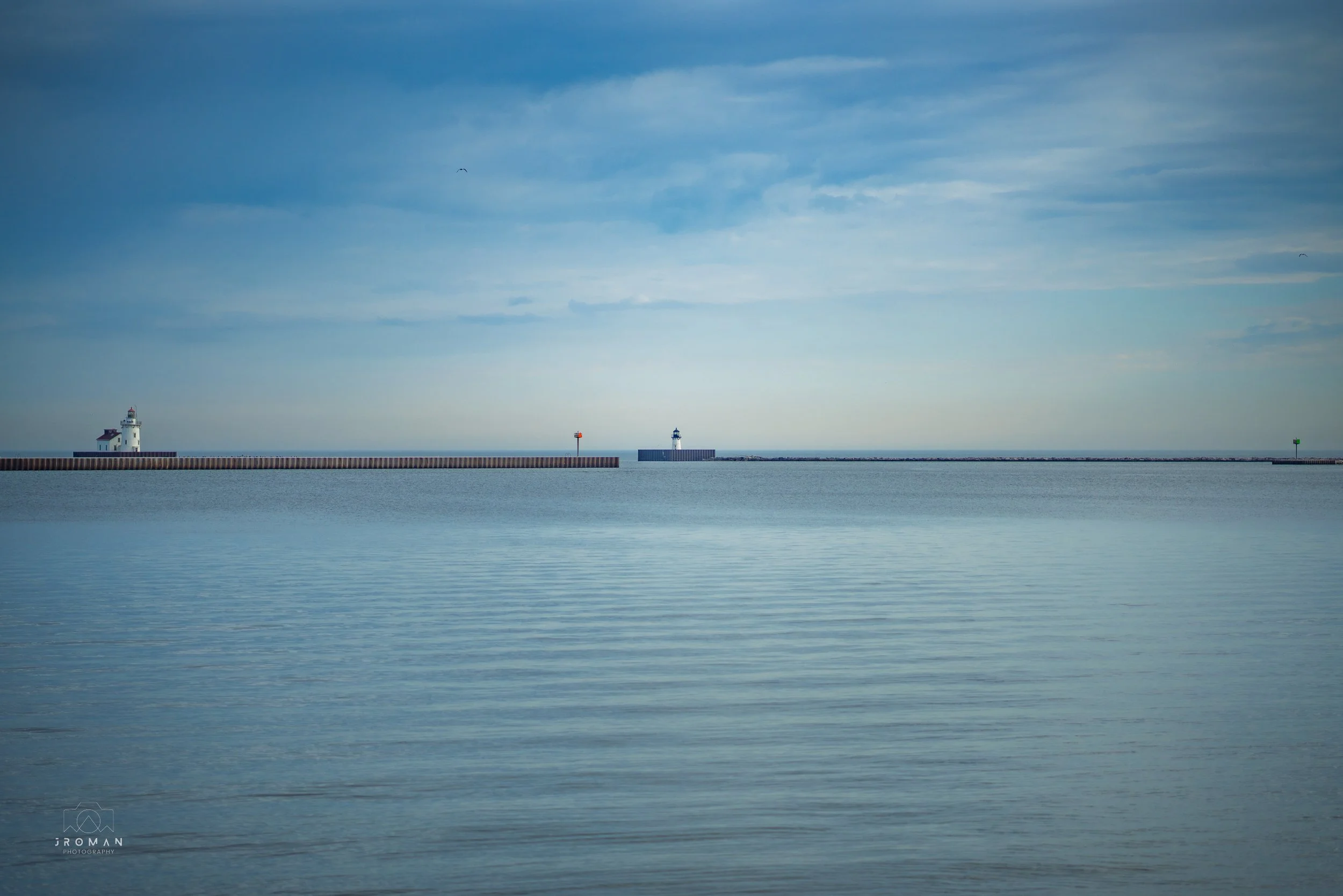 Calm sea with two lighthouses on breakwaters, one on the left and one in the center, under a cloudy sky.