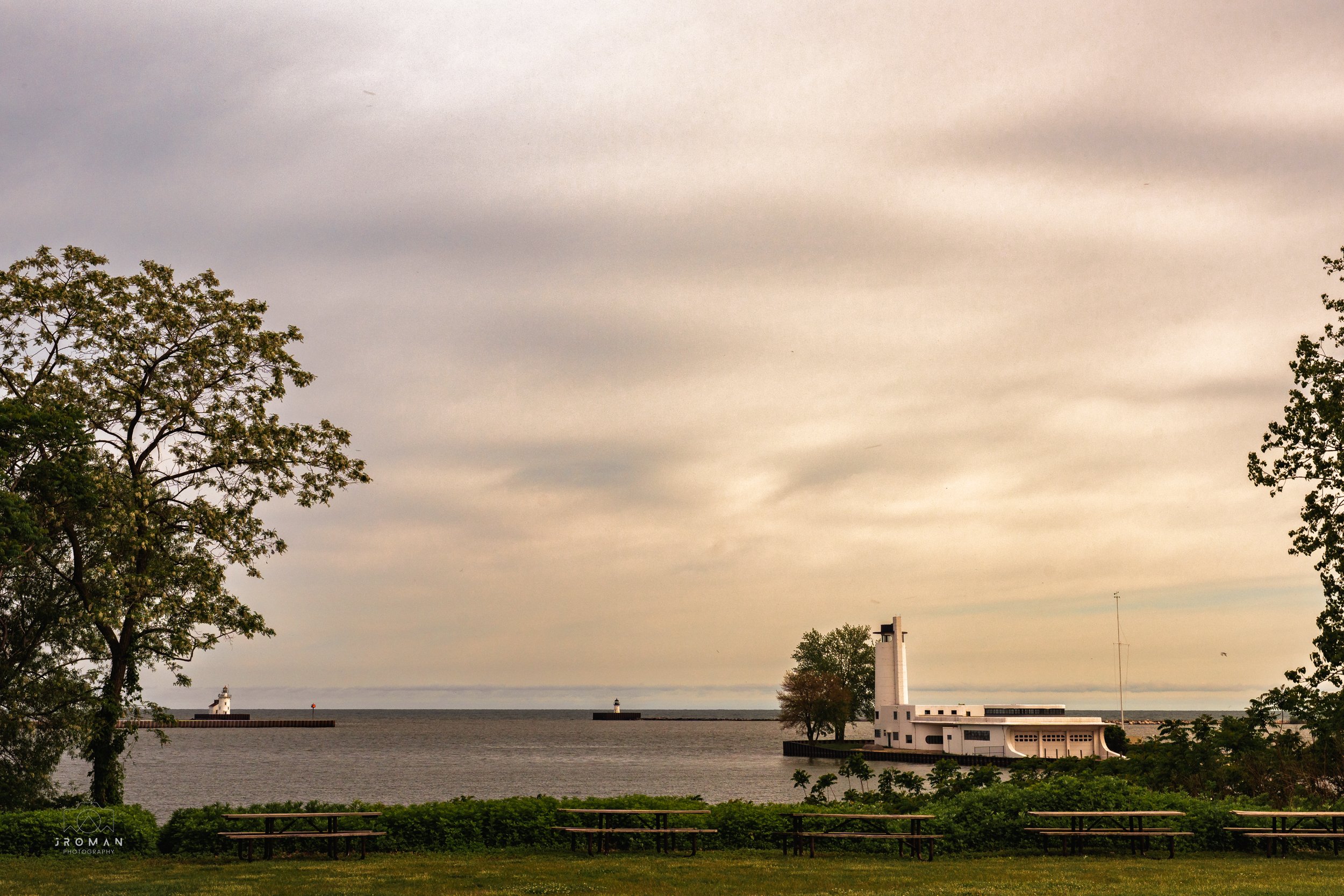 A scenic view of a waterfront with a lighthouse and a small white building, framed by trees and an overcast sky.