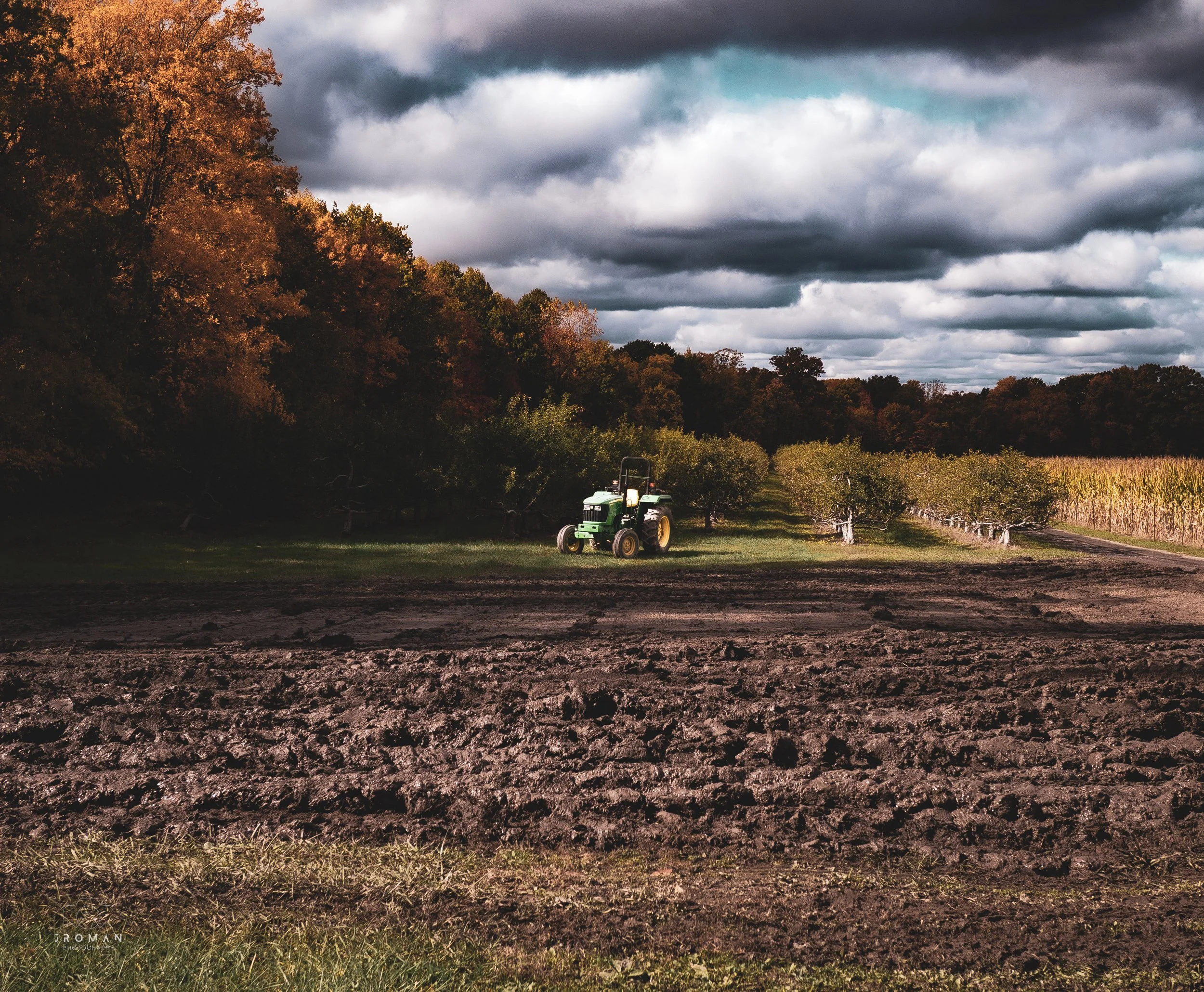 A green tractor on a farm field with rows of trees and an oak forest in the background. The sky is filled with dark, cloudy weather.