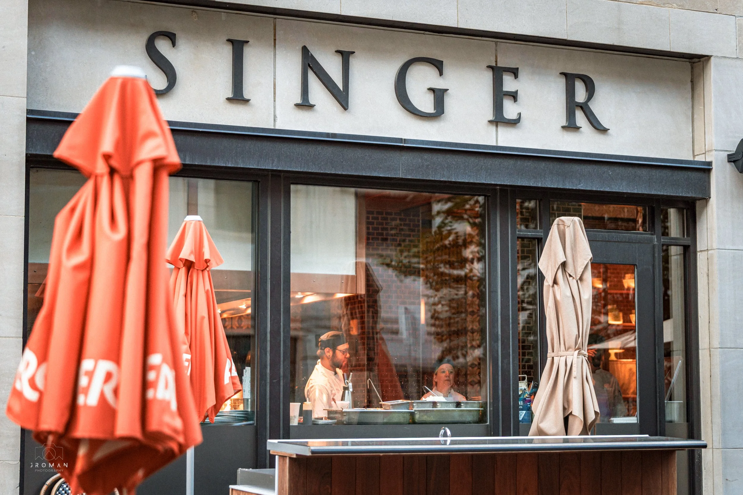 The exterior of a restaurant called SINGER with large black letters above the entrance, outdoor umbrellas, and a view into the kitchen through large windows showing chefs preparing food.