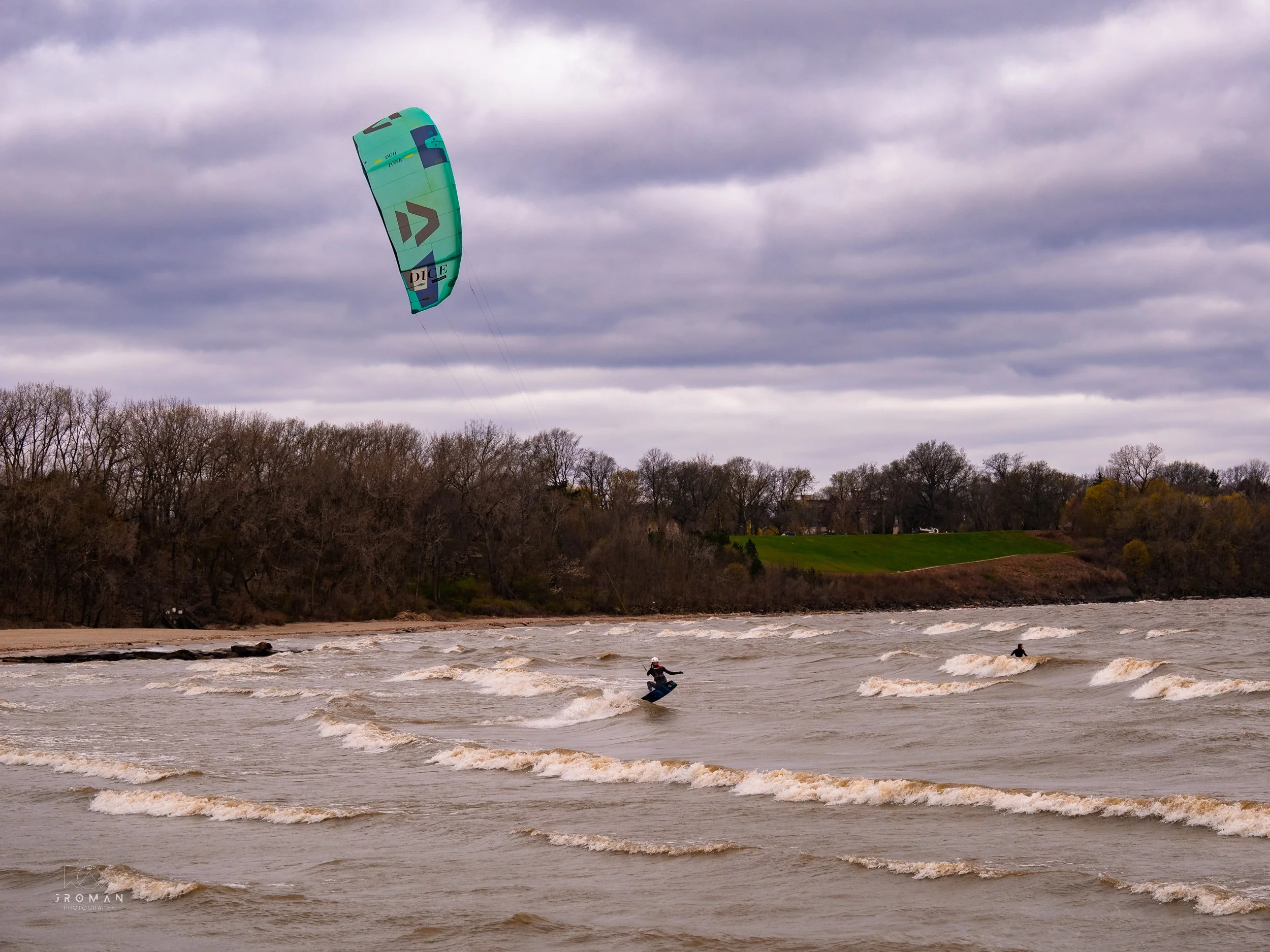 A person kiteboarding on a choppy river under a cloudy sky, with another person in the water and a tree-lined shoreline in the background.