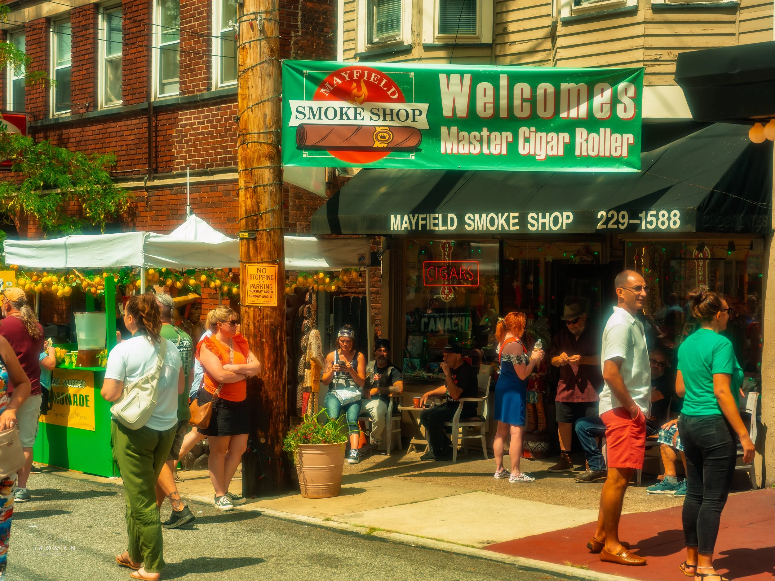 A busy outdoor street scene in front of the Mayfield Smoke Shop, with people gathered around outside and a green sign that reads 'Welcomes Master Cigar Roller'.