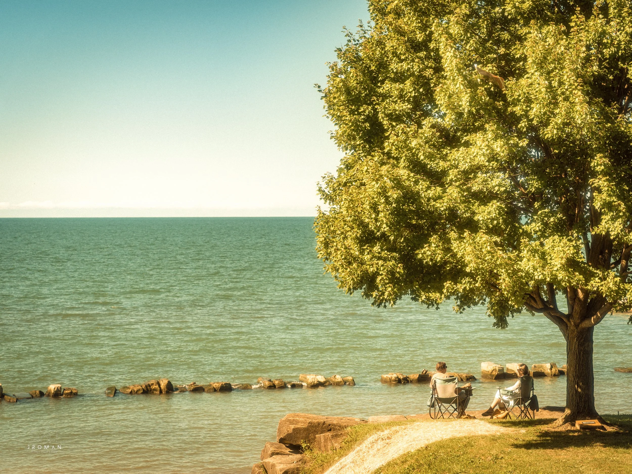 Two people sitting in camping chairs under a large green tree near the shoreline, with a calm body of water and a stone jetty or barriers extending into the water, on a sunny day.