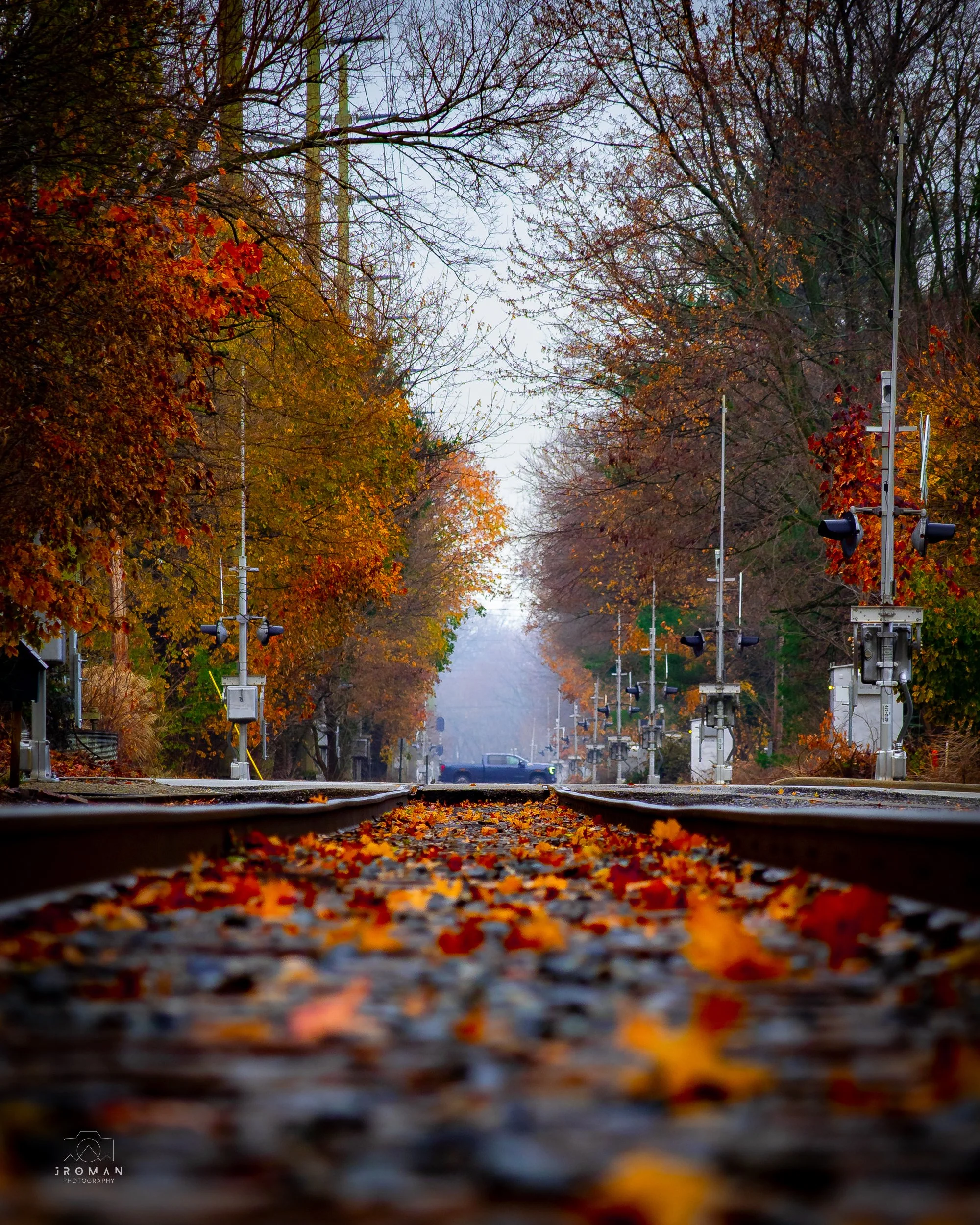 Photo of a railway track during autumn with fallen leaves scattered on the rails, surrounded by trees with orange and yellow foliage, and a traffic barrier in the distance.