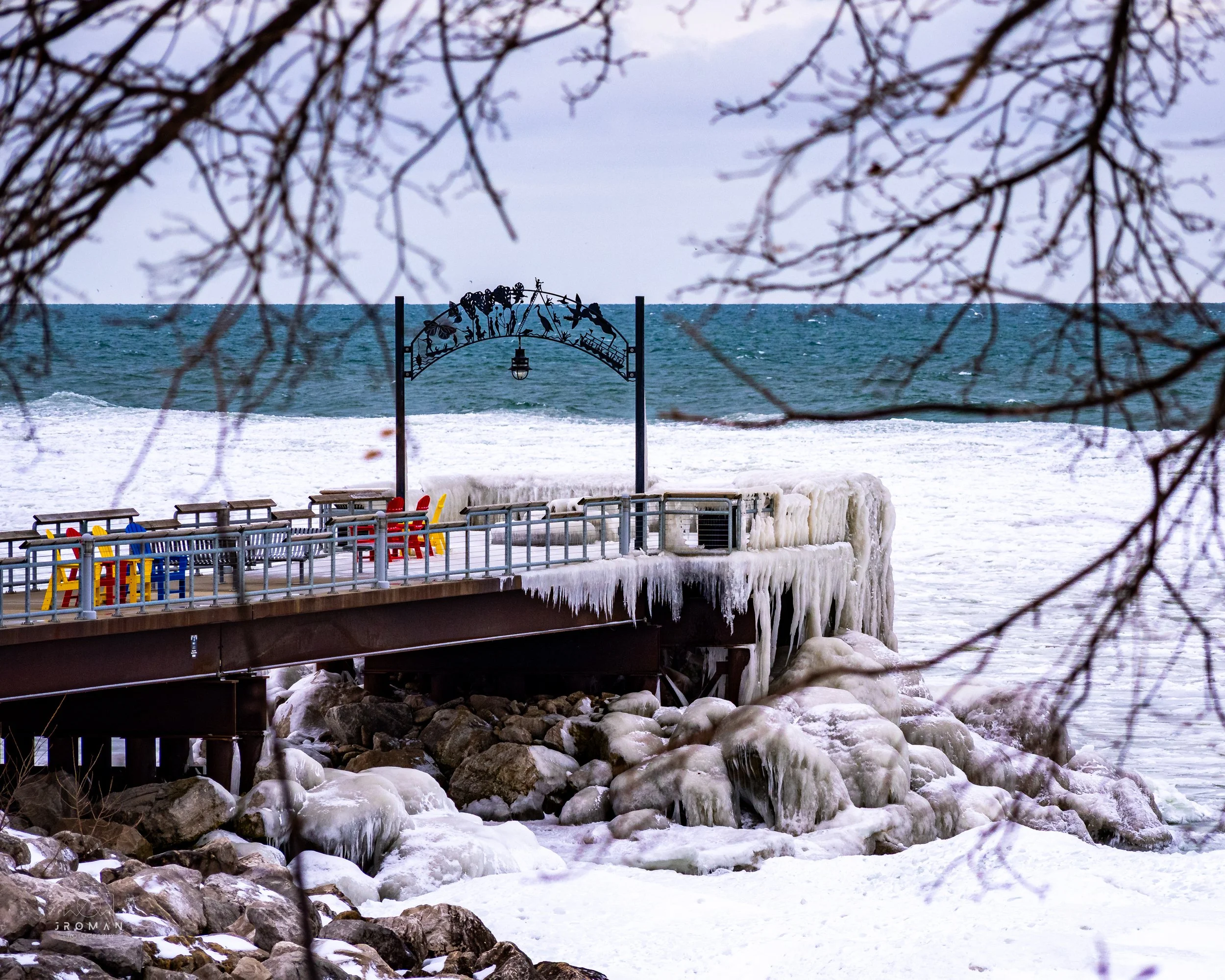 A frozen pier extends into the icy ocean, with colorful chairs on the deck, icicles hanging from the railings, and snow-covered rocks underneath. Bare tree branches frame the scene.