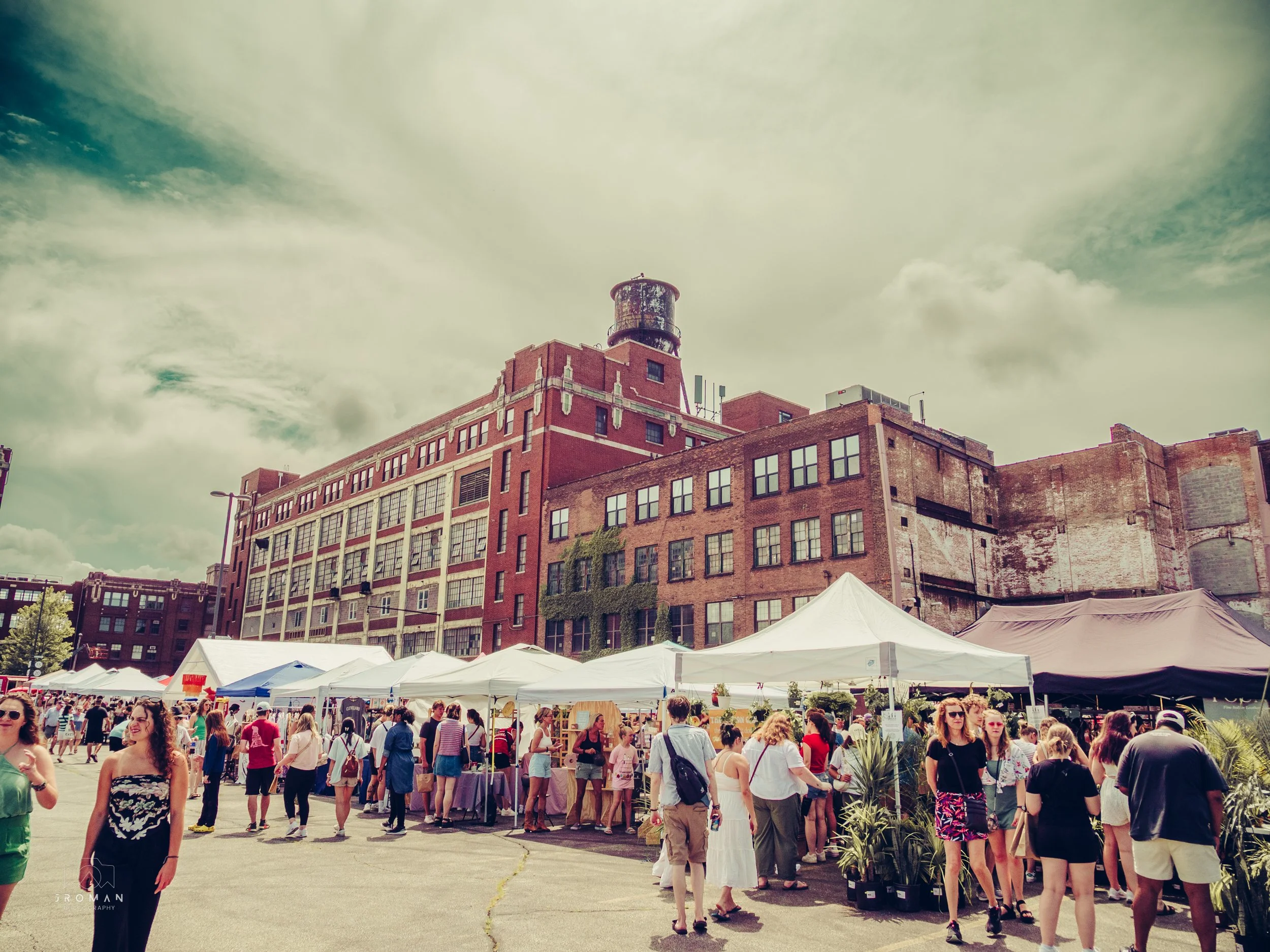 Outdoor market with white tents and people shopping in front of a large brick building under a partly cloudy sky.
