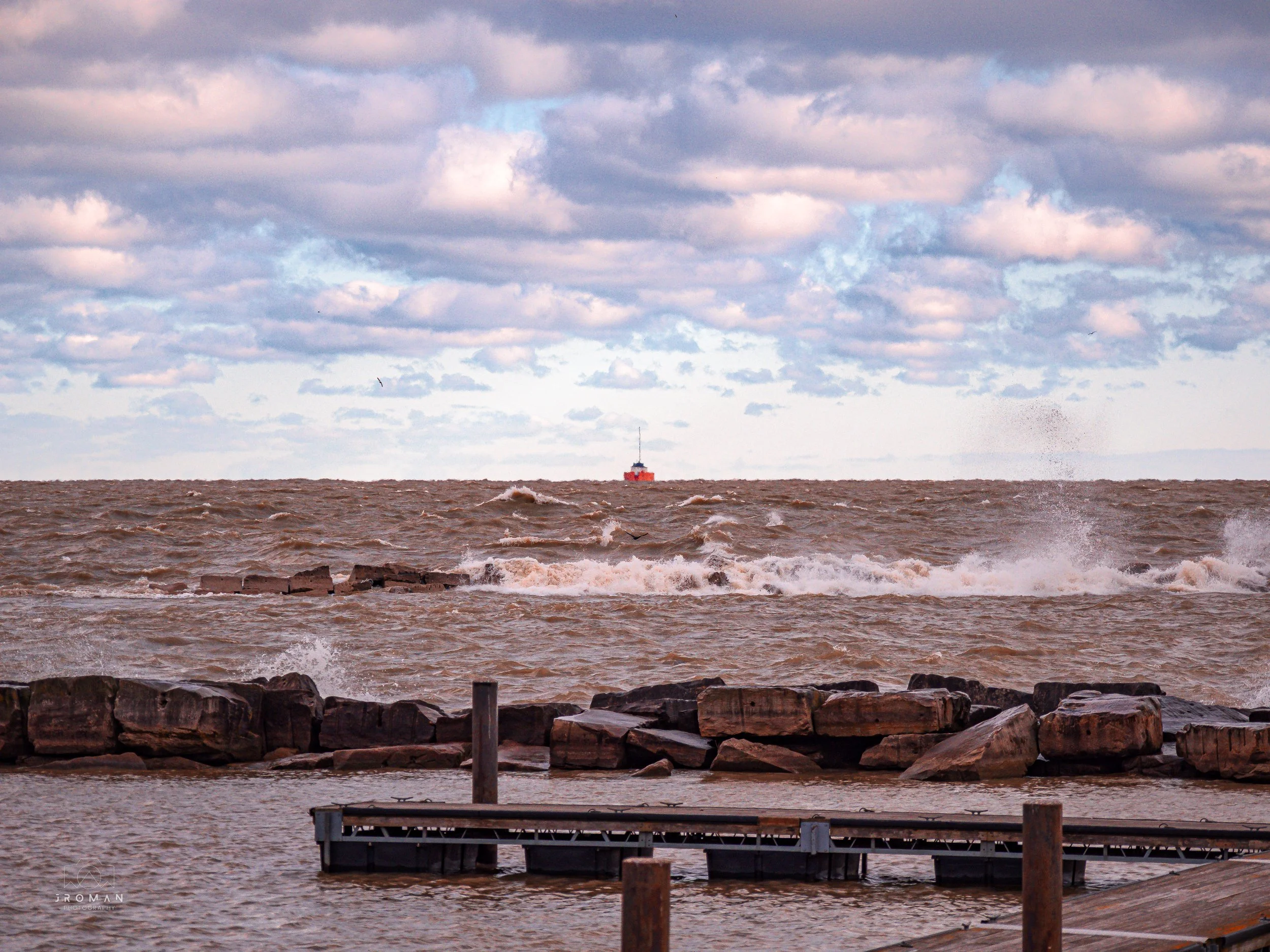 A photo of a body of water with waves crashing against rocks, a dock in the foreground, and a boat in the distance under a cloudy sky.