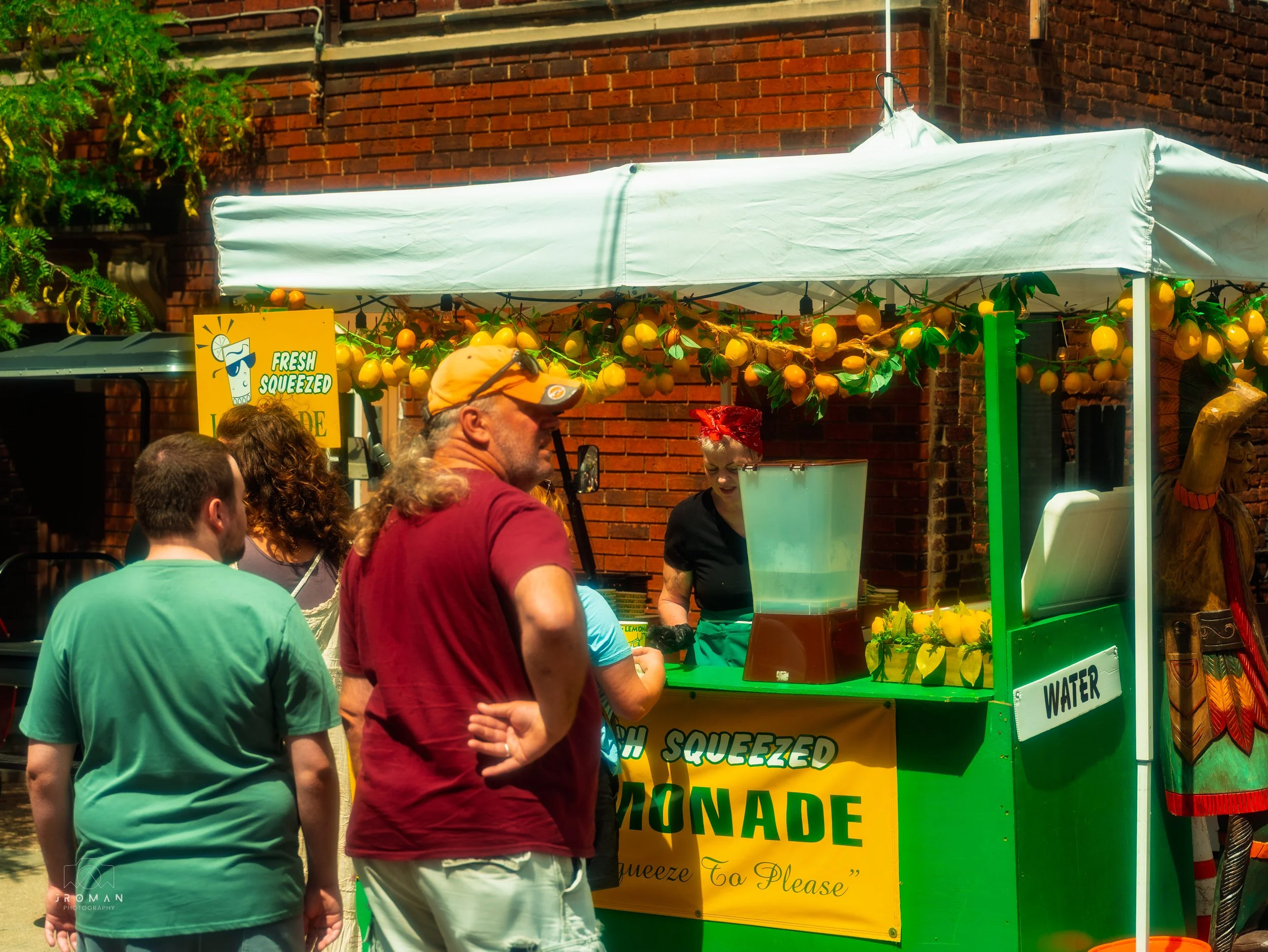 People standing in line at a lemon juice stand with a sign that says 'Fresh Squeezed Lemonade,' decorated with hanging lemons and greenery, on a sunny day.