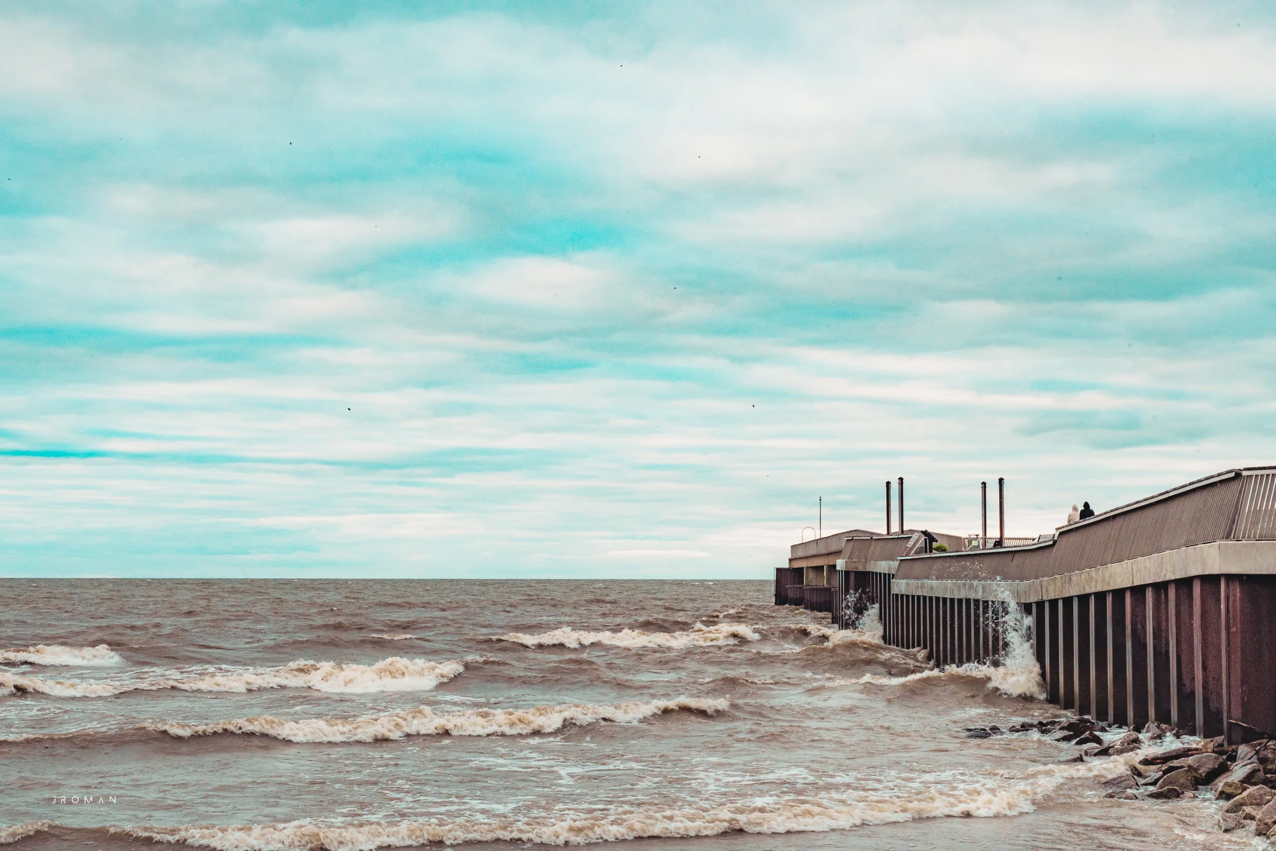 Overcast sky with waves crashing against a sea wall or pier extending into a turbulent ocean.