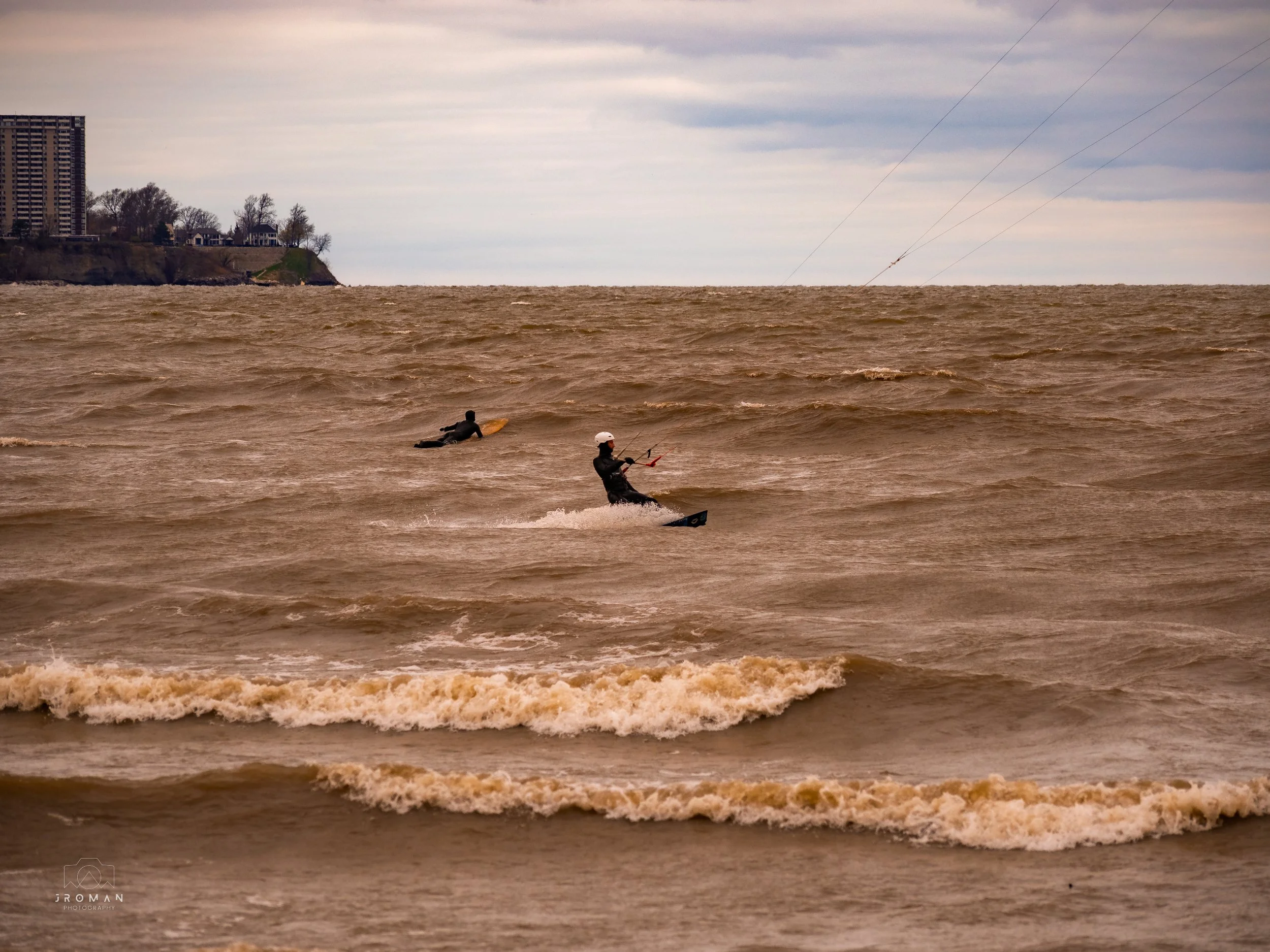 Two people kite surfing on choppy brown water near a beach during cloudy weather, with a cityscape in the background.