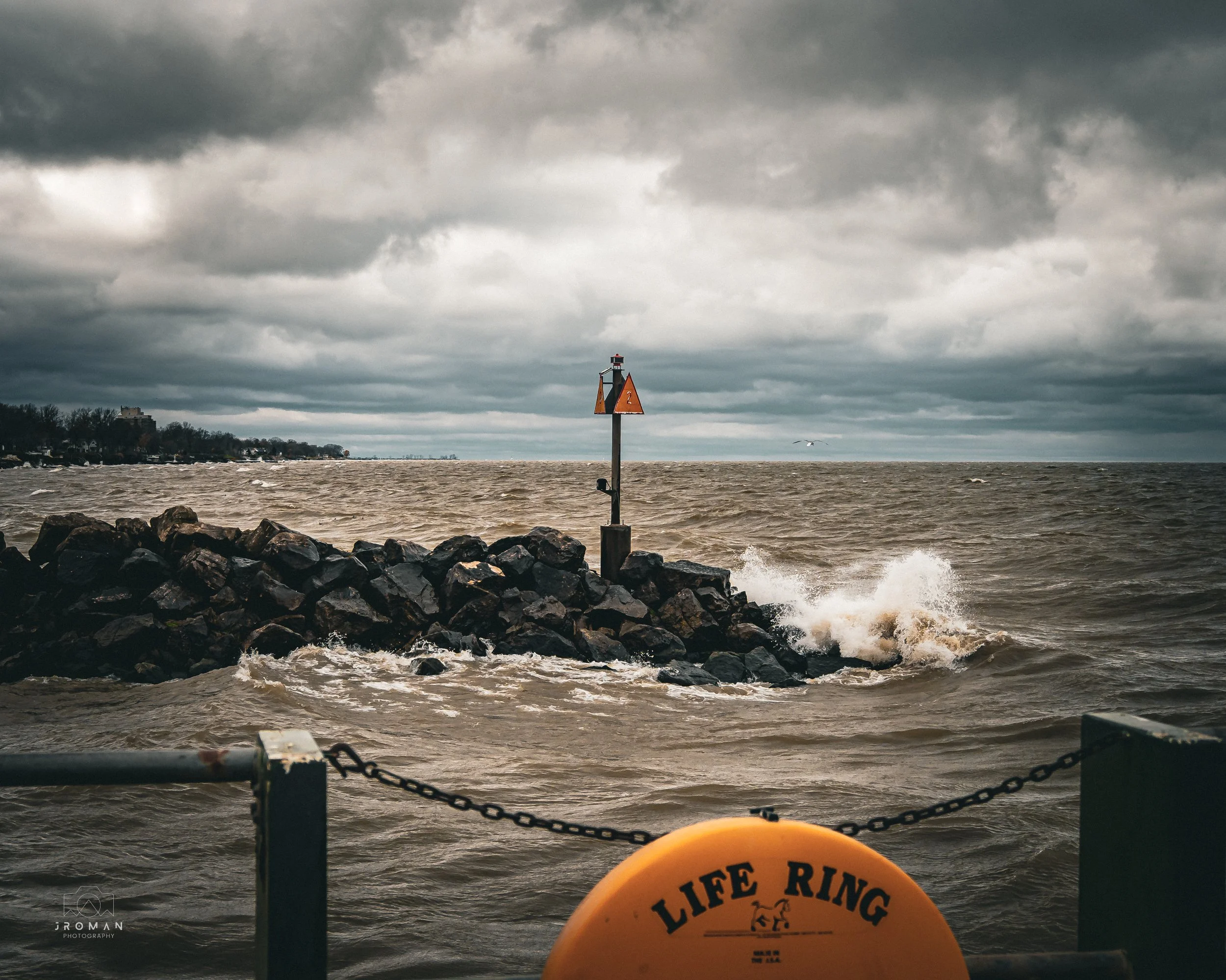 A stormy scene at a body of water, with dark clouds overhead, waves crashing against a rocky breakwater, a small lighthouse on a pole, and a yellow life ring in the foreground.