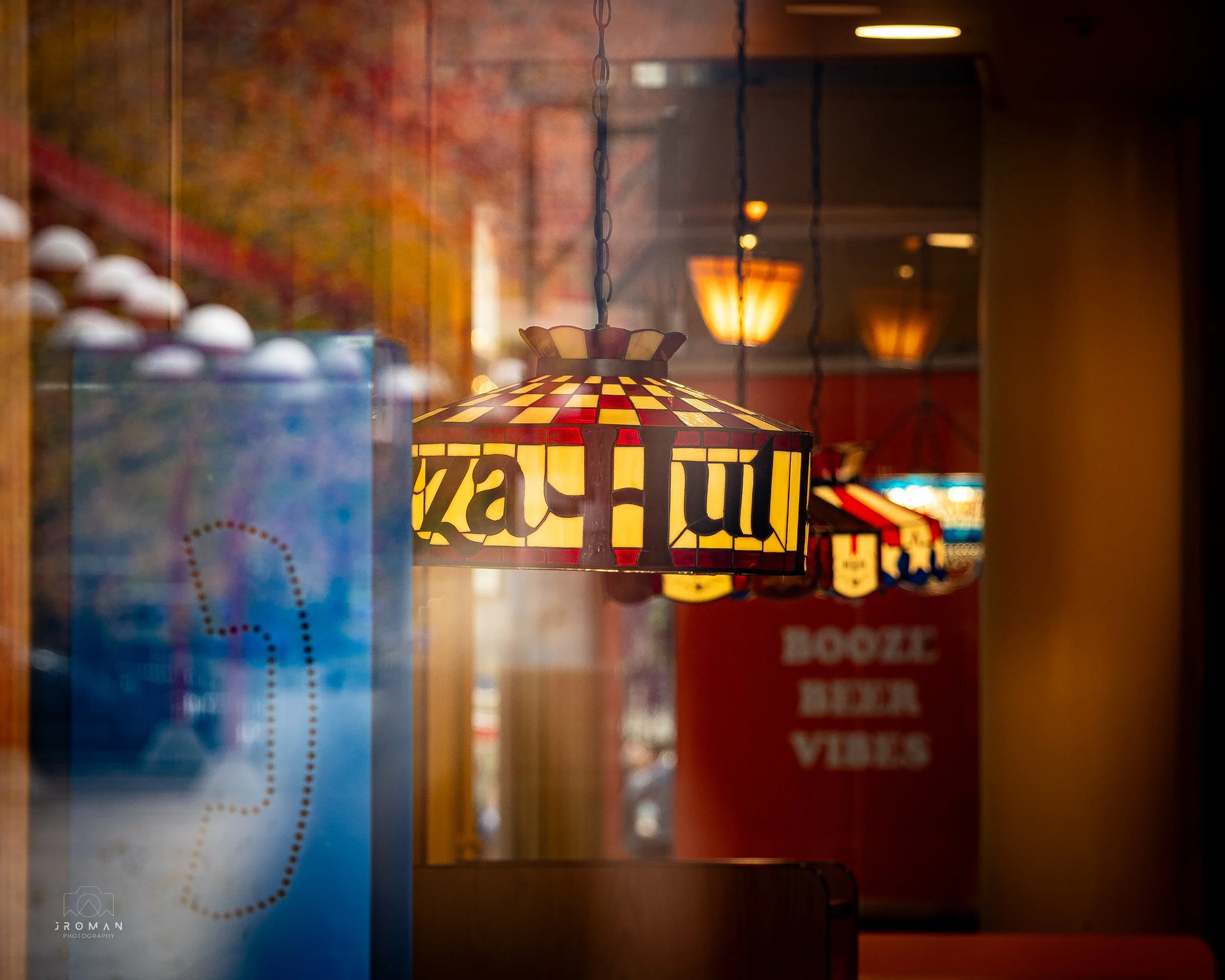 Decorative stained glass hanging lamp inside a restaurant or bar with warm lighting, with a blurry background showing a sign that says 'Booze Beer Vibes' and some colorful lights.