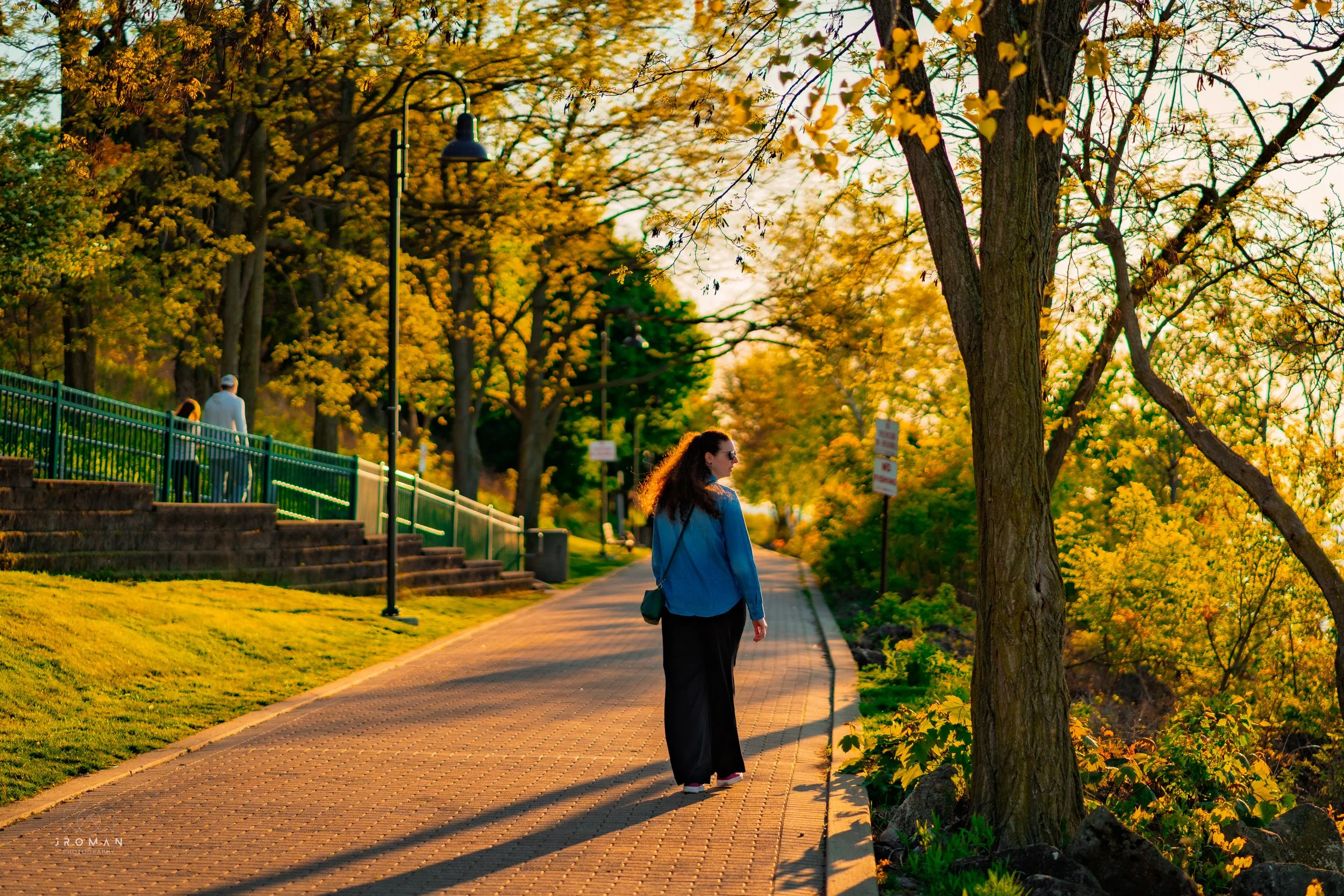 A woman with curly hair wearing sunglasses, a blue jacket, and black pants, walks along a paved pathway in a park surrounded by trees with yellow and green leaves during sunset.