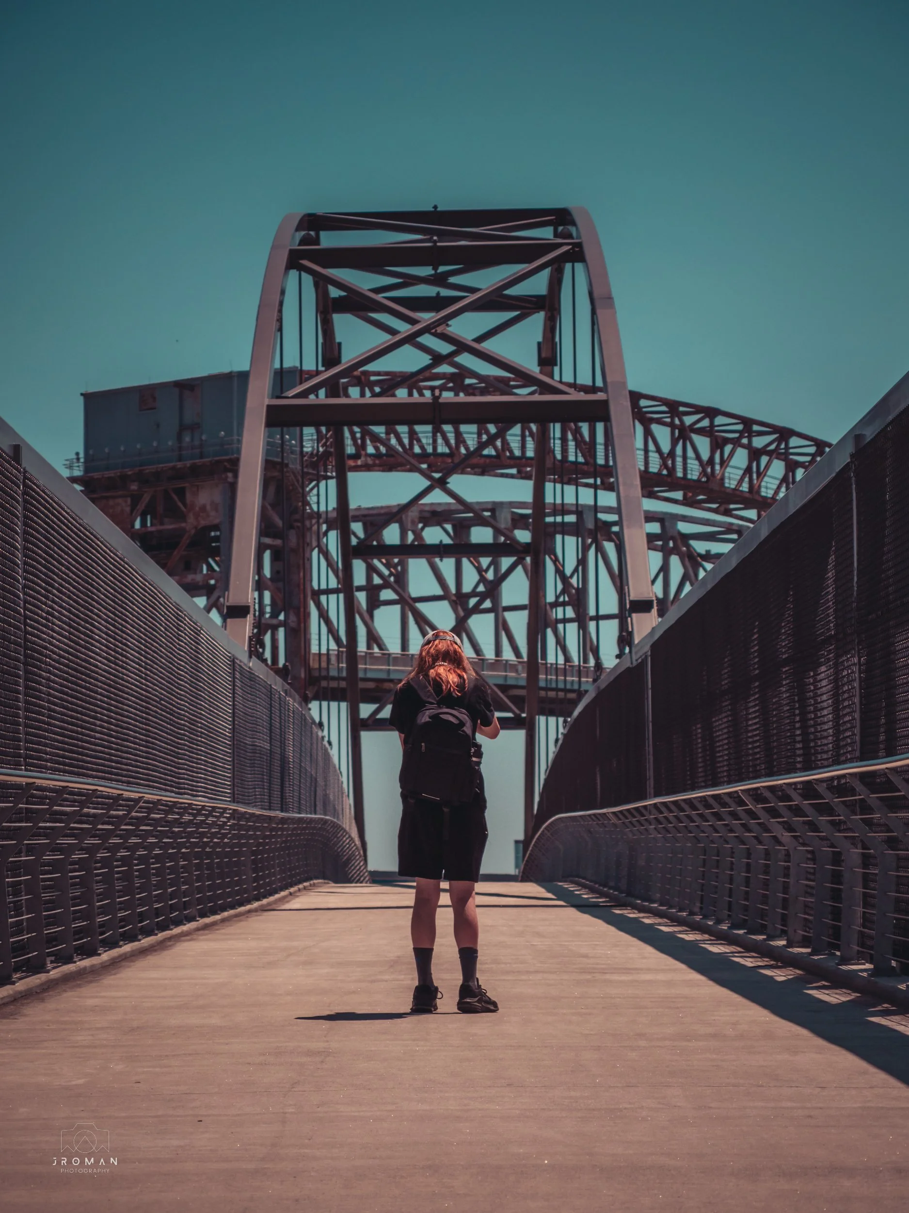 A person standing on a bridge with a backpack, looking up at a large industrial structure and arching metal framework against a blue sky.