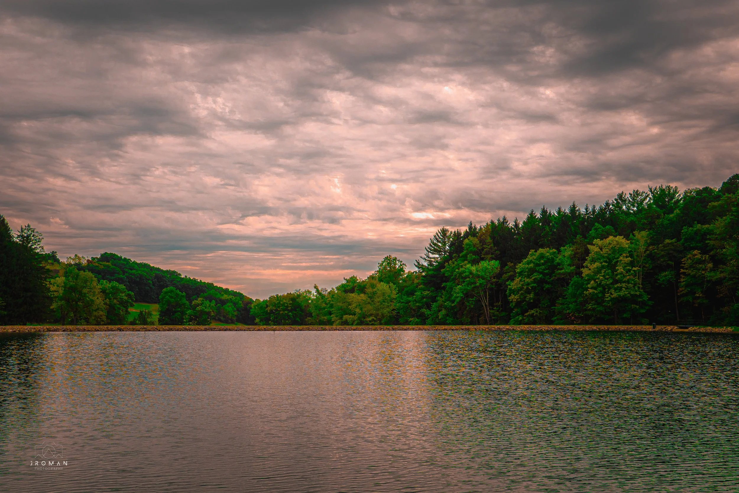 A peaceful lake surrounded by lush green trees with a cloudy sky overhead, reflecting on the water surface.