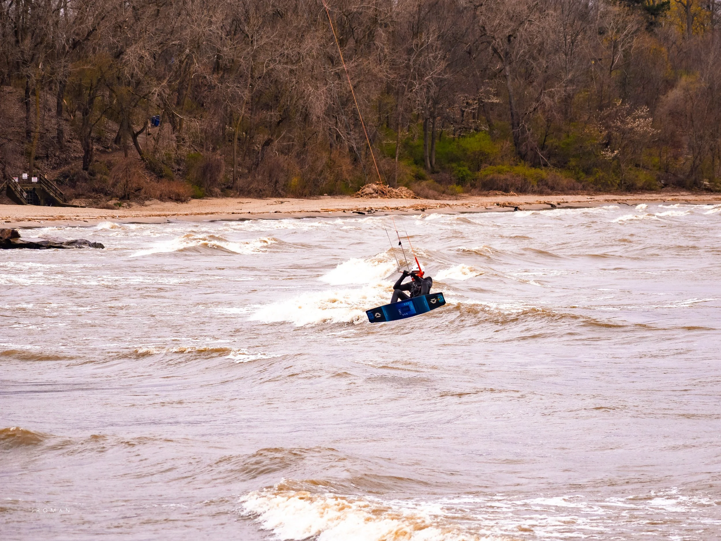 Person in a wetsuit kiteboarding on a choppy body of water near a sandy beach with trees in the background.