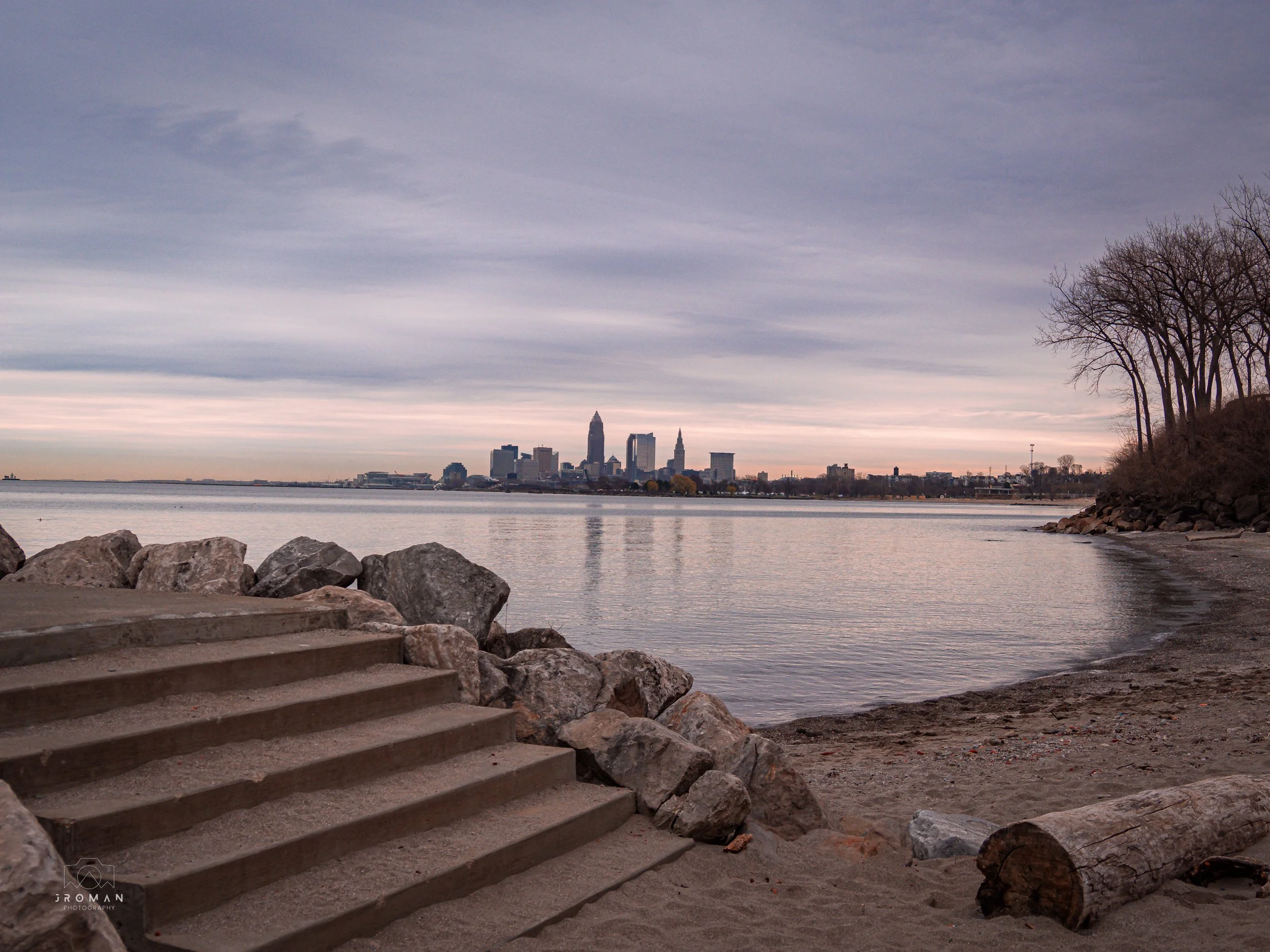 View of Cleveland skyline from the lakeshore with stone stairs and logs in the foreground, and leafless trees on a small hill on the right.