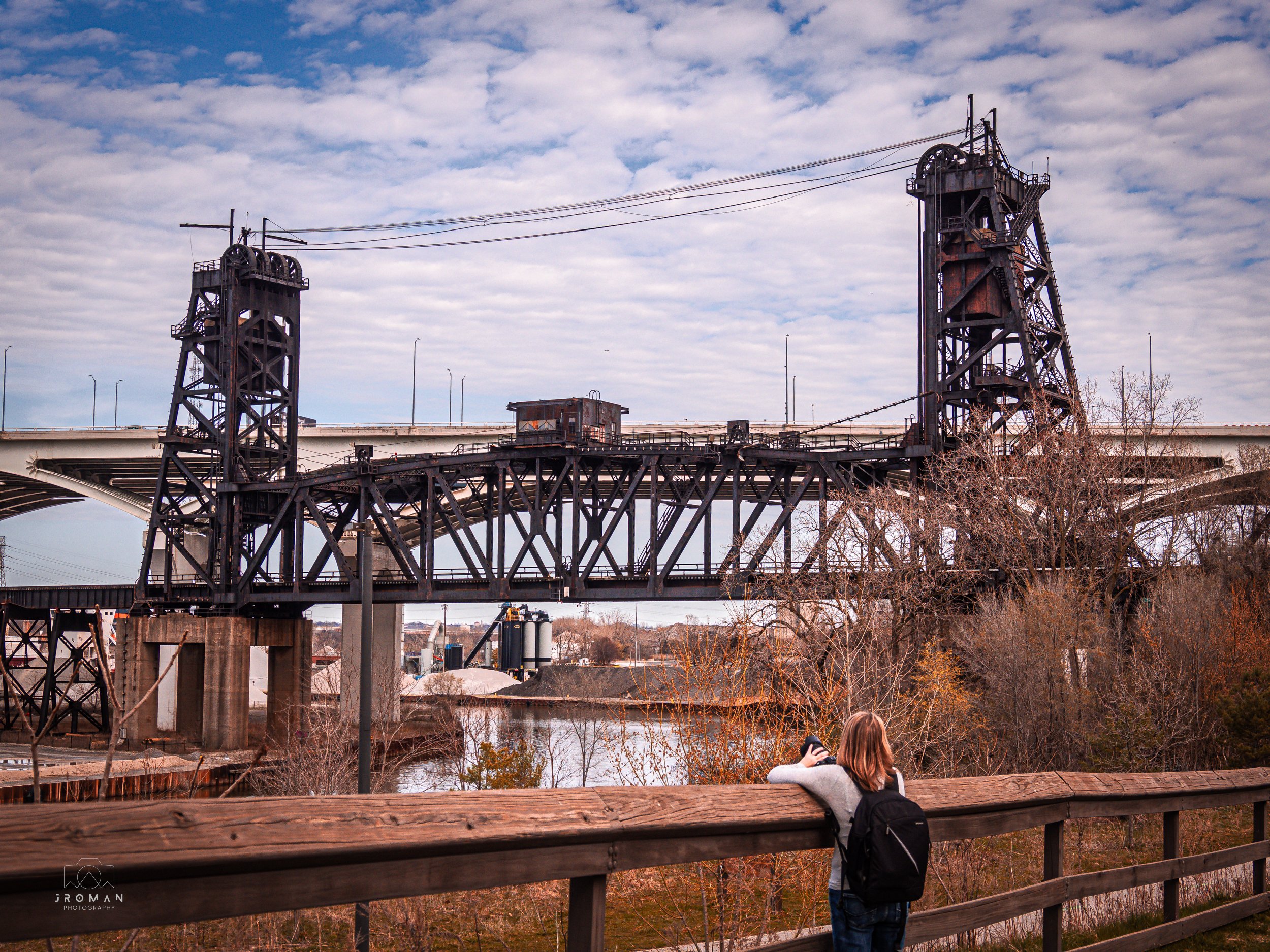 A woman with a backpack leaning on a wooden railing, gazing at an old industrial bridge with two tall towers over a river, under a cloudy sky.