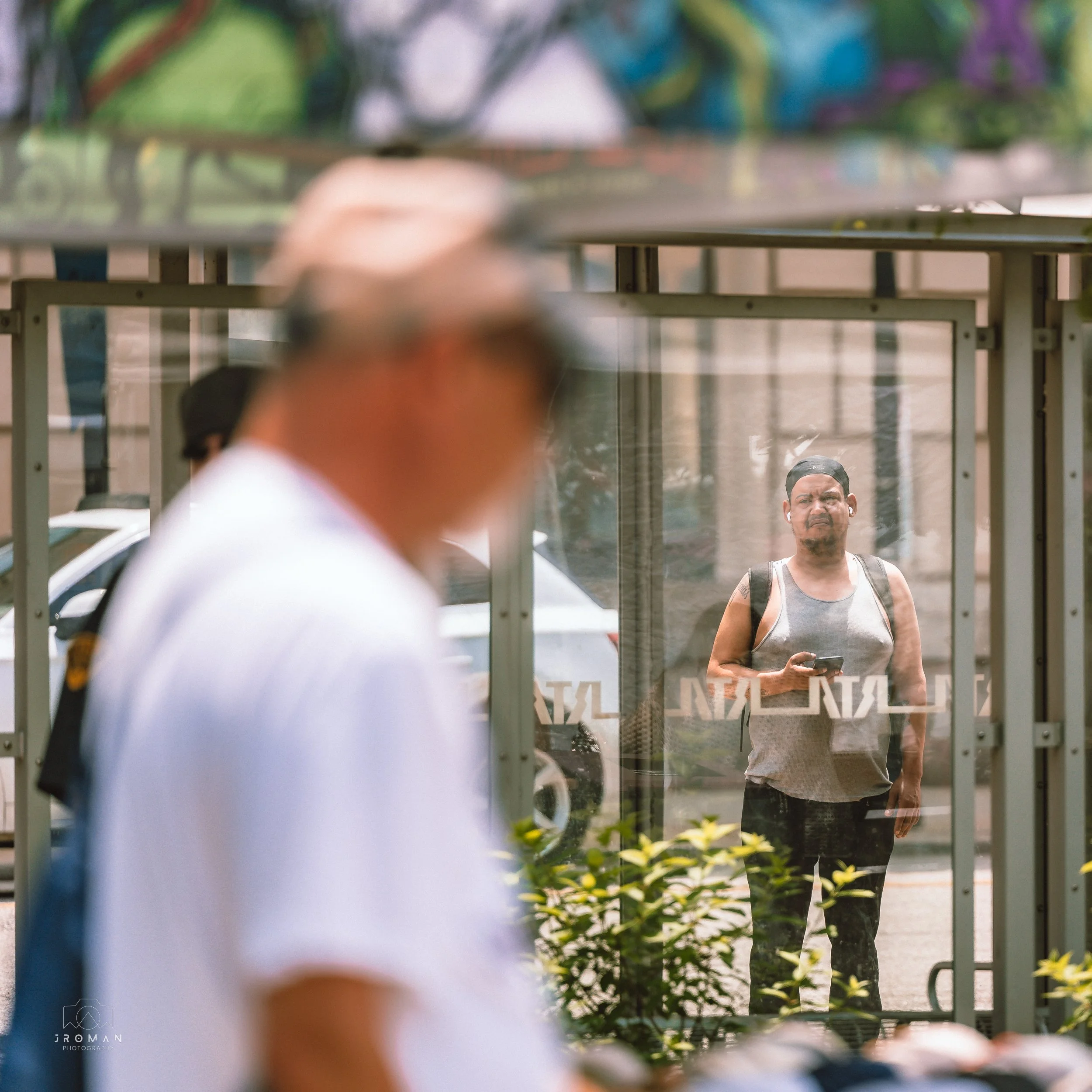 A man with a backpack and a smartphone looking through a glass window at a blurred person passing by in the foreground.