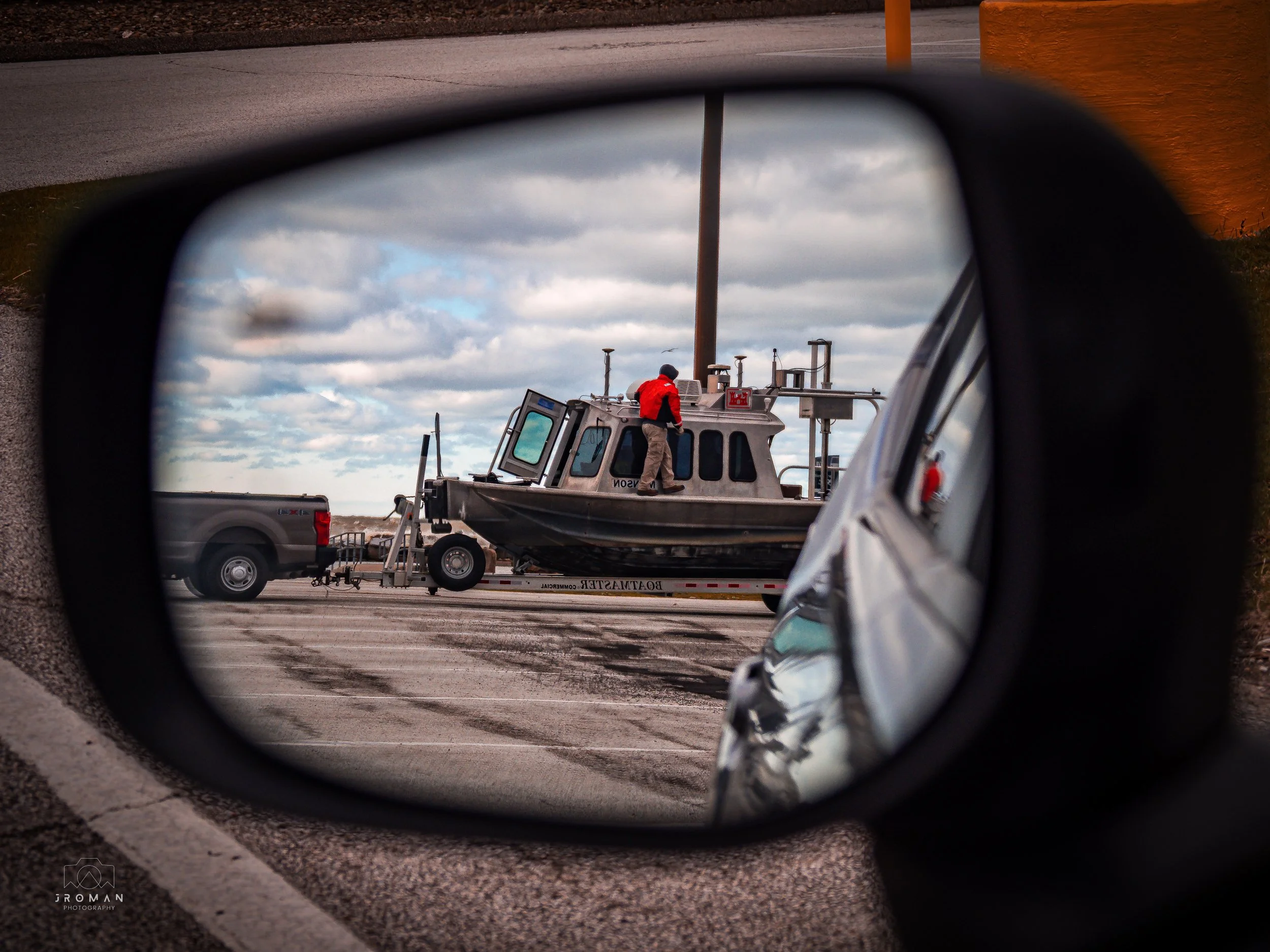 Person in red jacket on top of a boat on a trailer, viewed through a car side mirror, with a cloudy sky in the background.