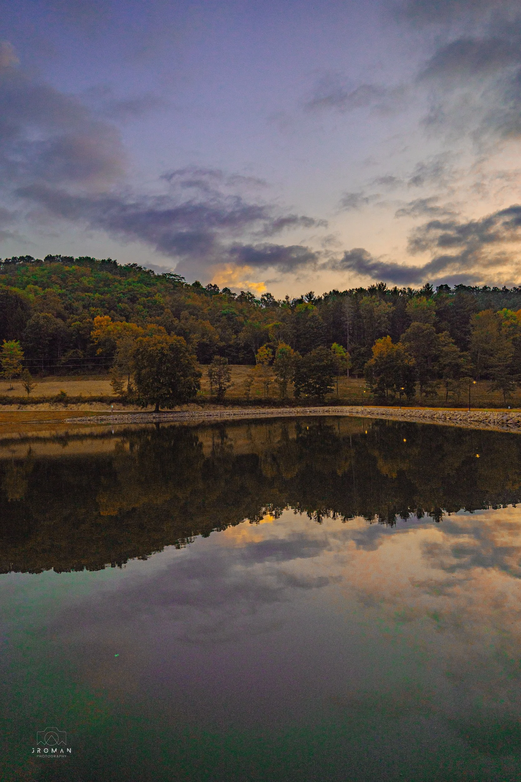 A landscape scene at dusk showing a lake with a reflection of the sky, clouds, and trees on its surface, with a wooded hillside in the background.