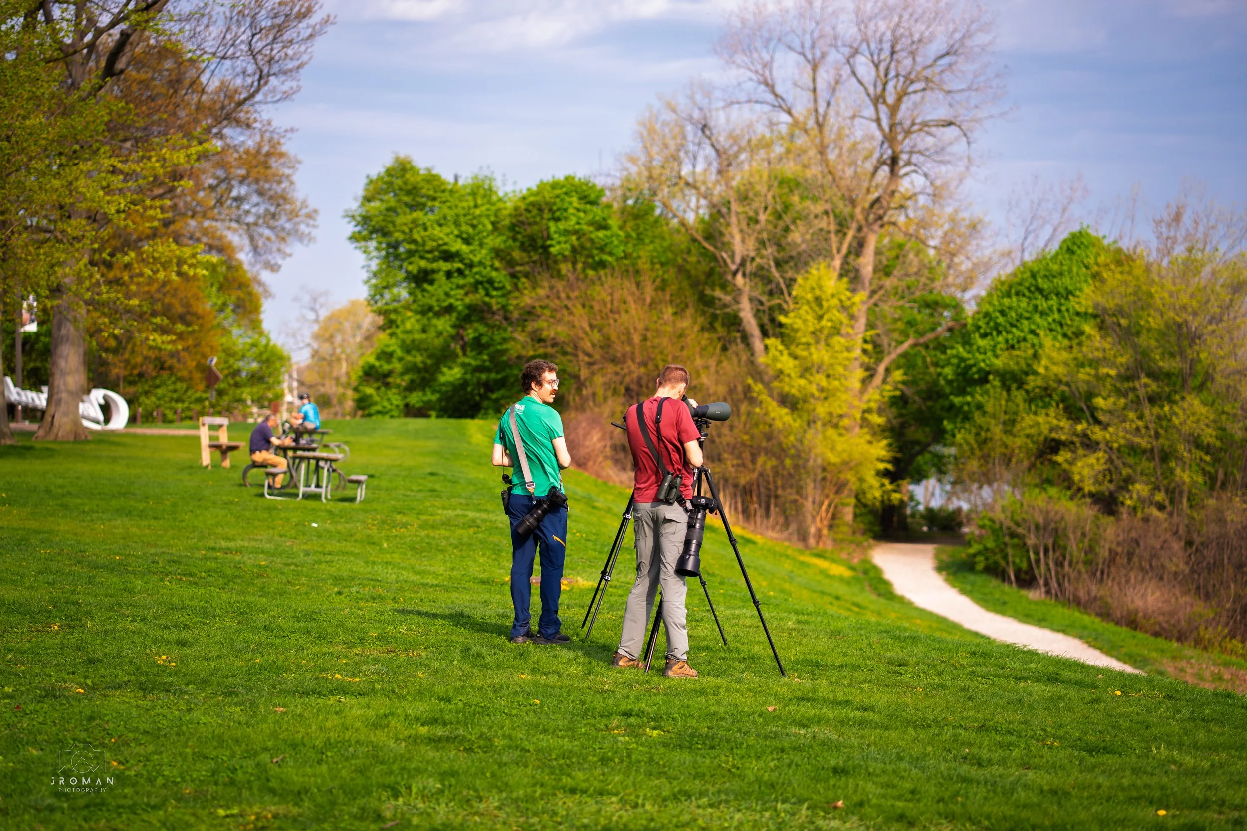Two photographers with cameras on tripods capturing a scenic landscape in a park with green grass, trees, and a walking path, while three people sit on benches nearby.