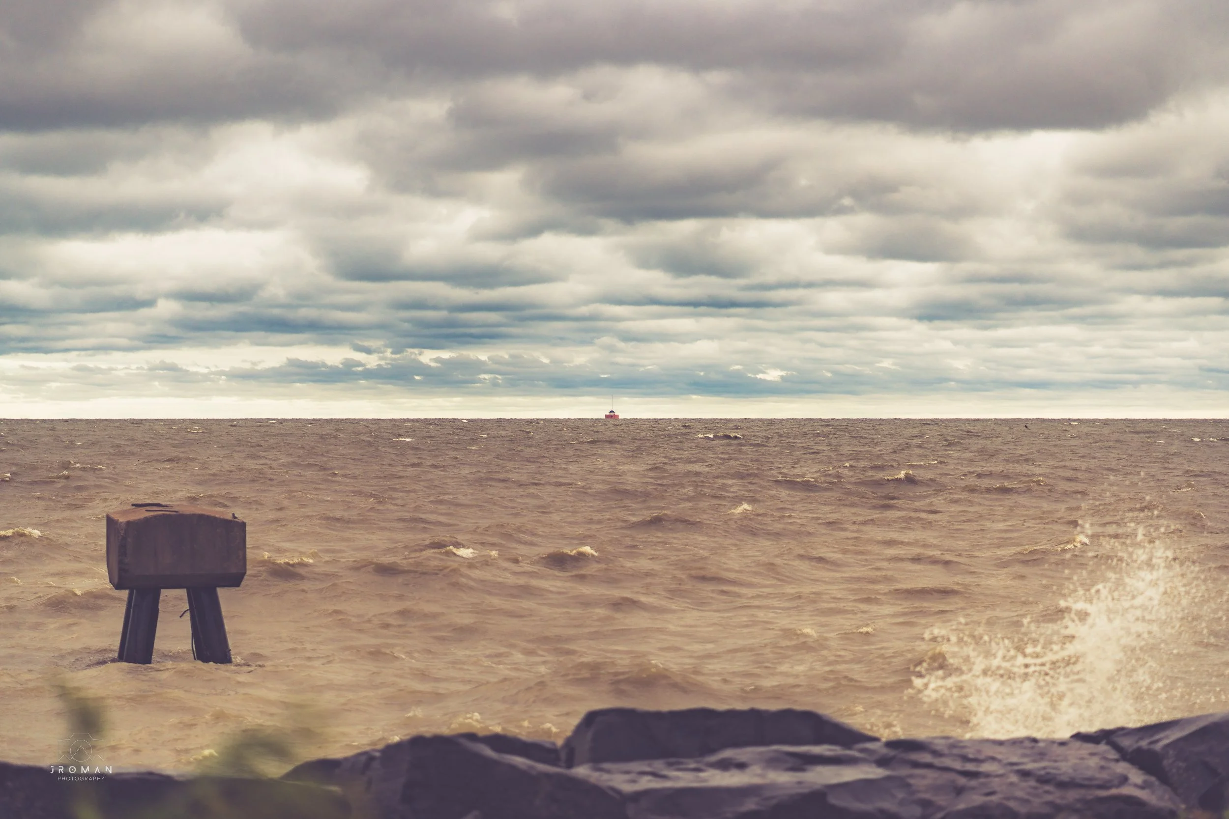 A stormy ocean scene with choppy waves, a dark cloudy sky, a small structure in the water, and a ship in the distance on the horizon.