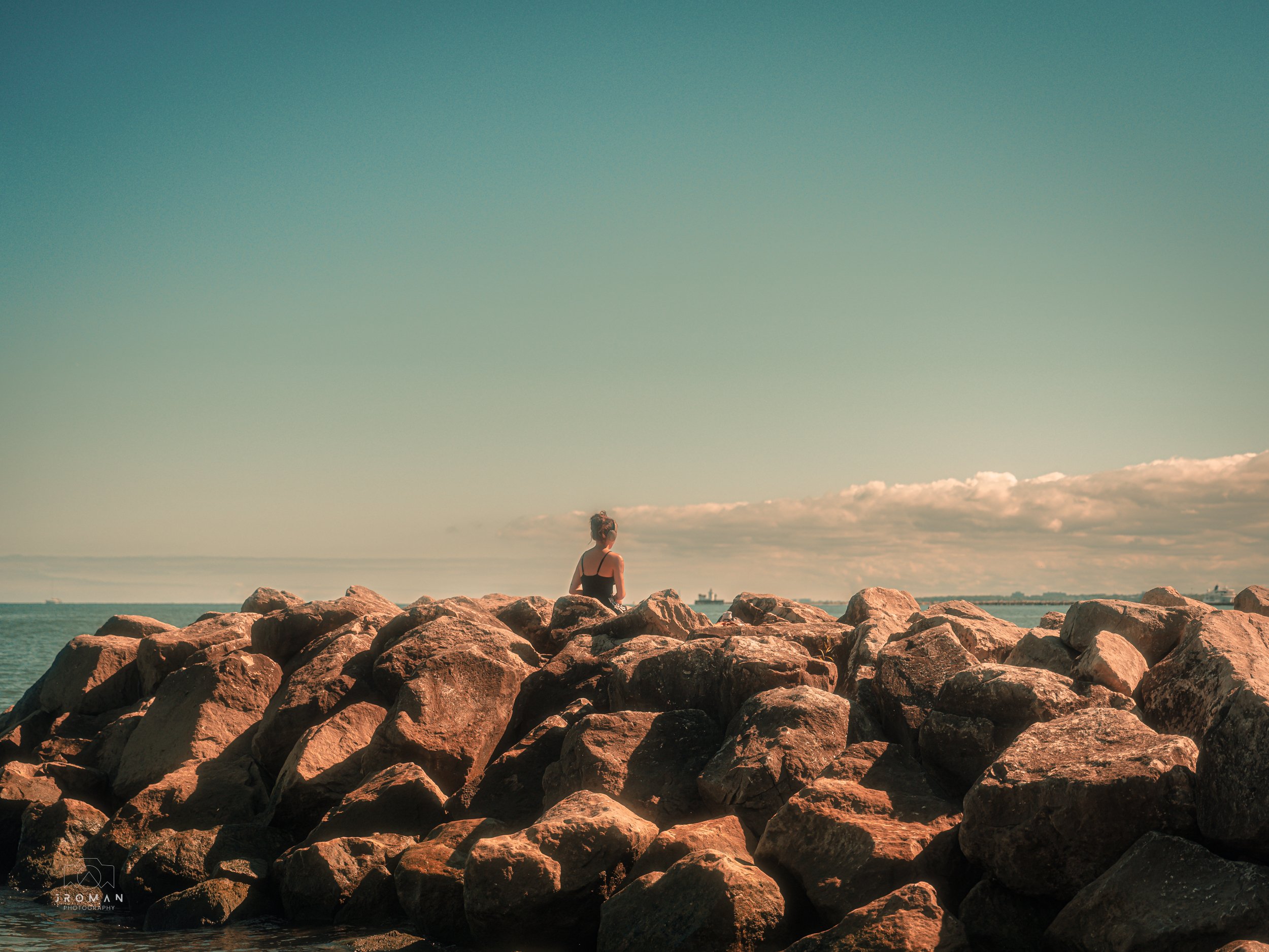 A woman sitting on large rocks by the ocean, facing away, under a mostly clear sky with a few clouds.