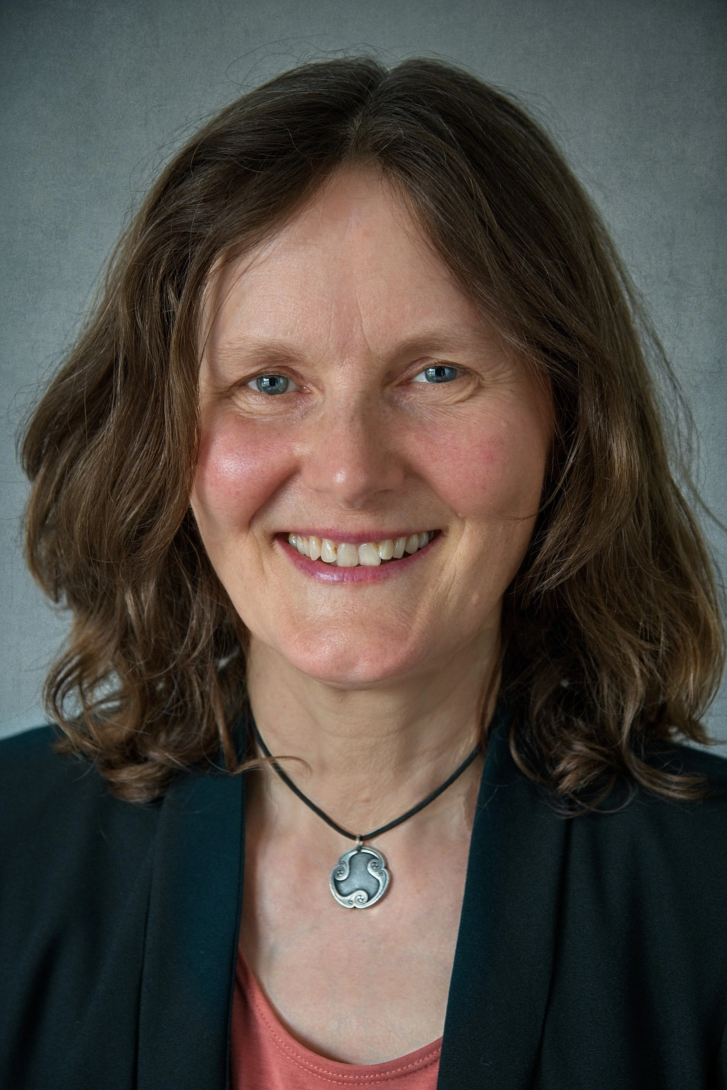 A woman with shoulder-length brown hair, blue eyes, and a friendly smile wearing a black blazer and a circular pendant necklace.