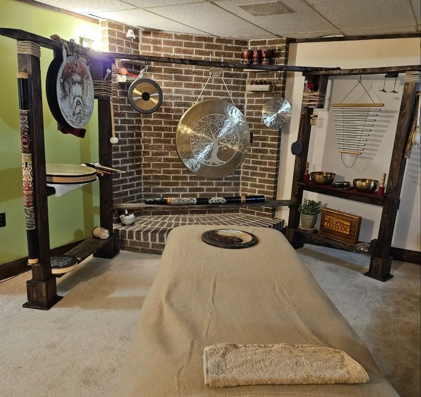 Room with a massage table covered in a beige sheet, surrounded by musical and meditation instruments including gongs, bowls, and a chime, with a brick wall and shelves in the background.