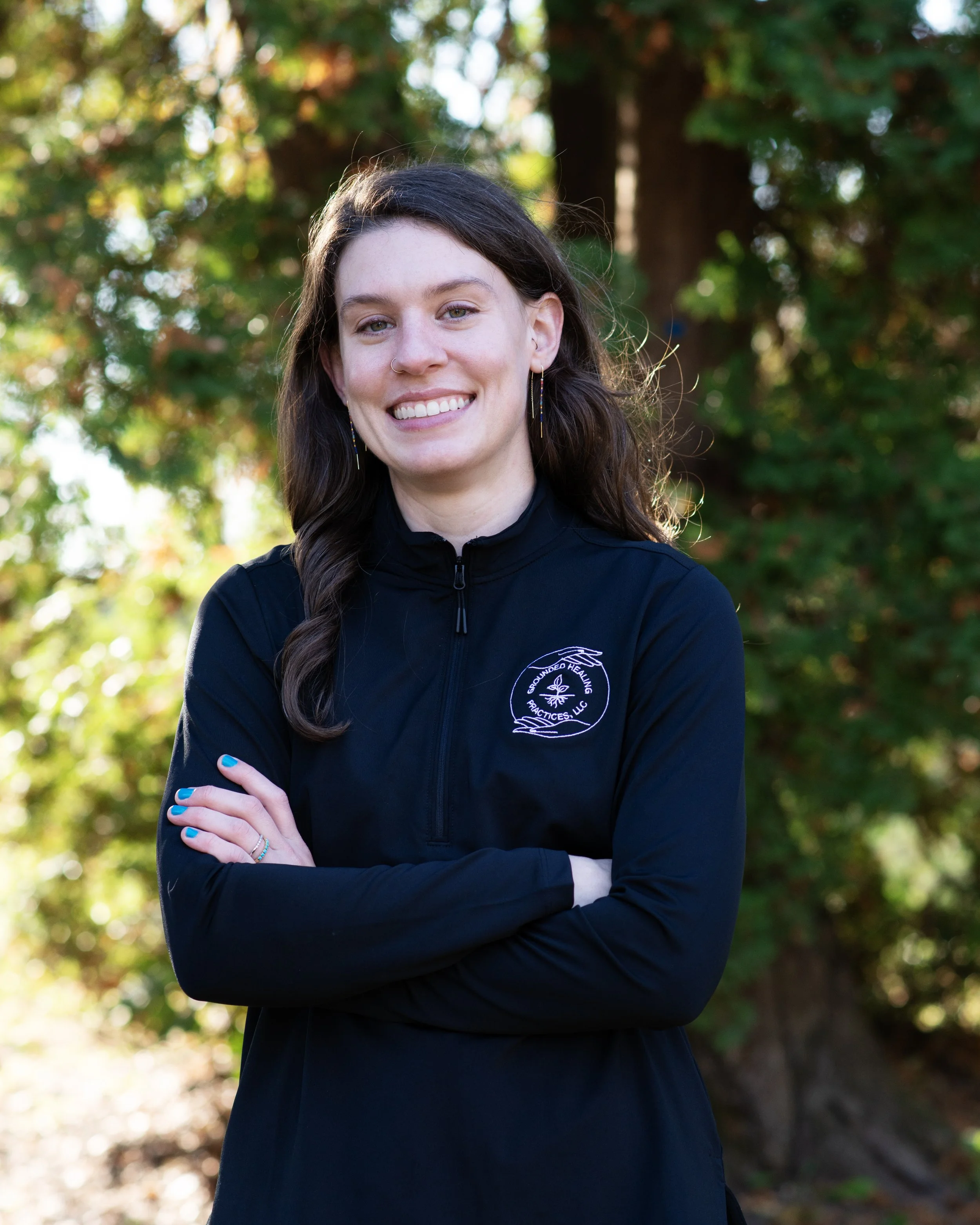 A smiling young woman with dark hair, wearing earrings and a black jacket with a white logo, standing outdoors with trees and foliage in the background.