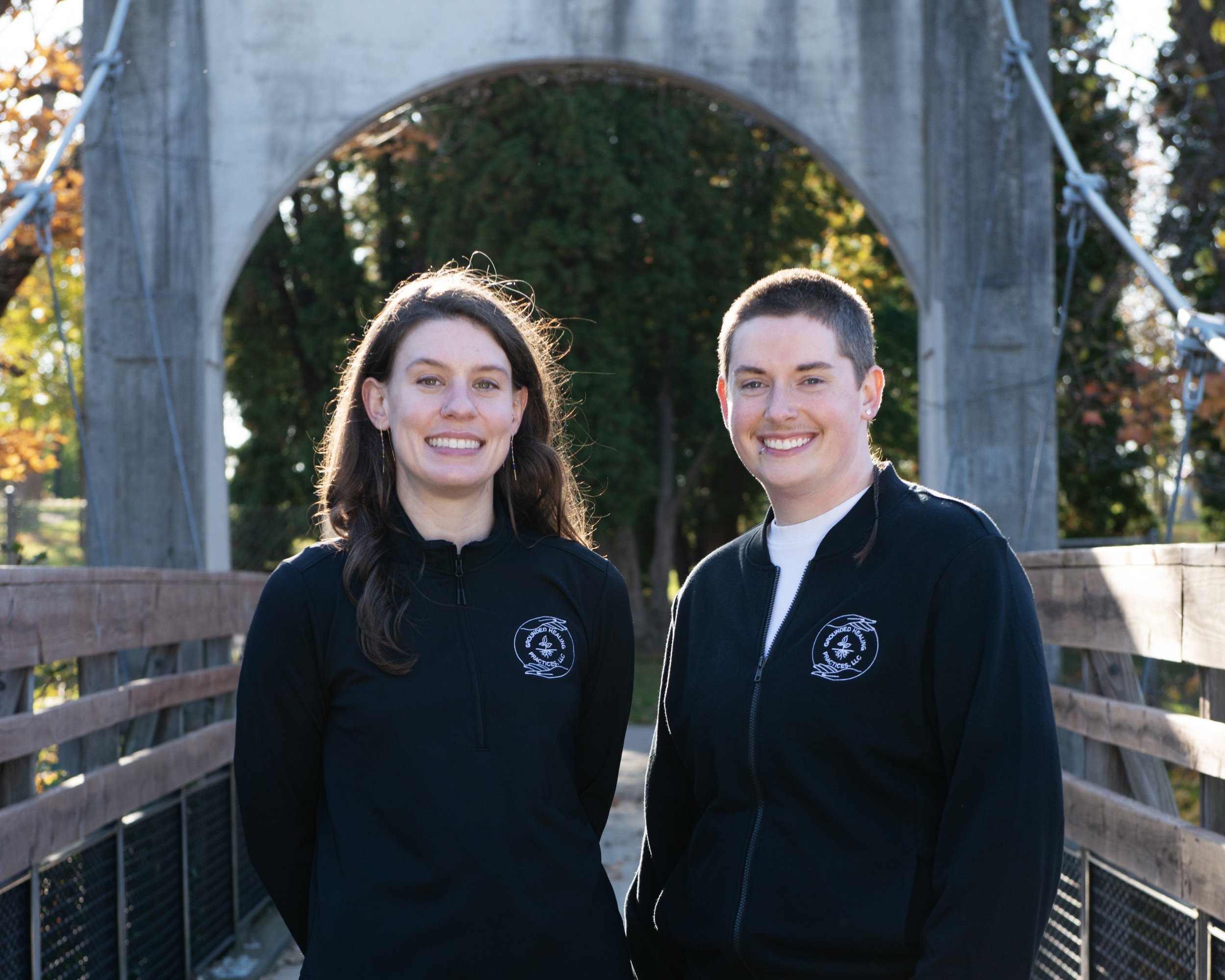 Two smiling women standing on a bridge in a park with trees and autumn leaves in the background, both wearing black jackets with a logo on the left side.