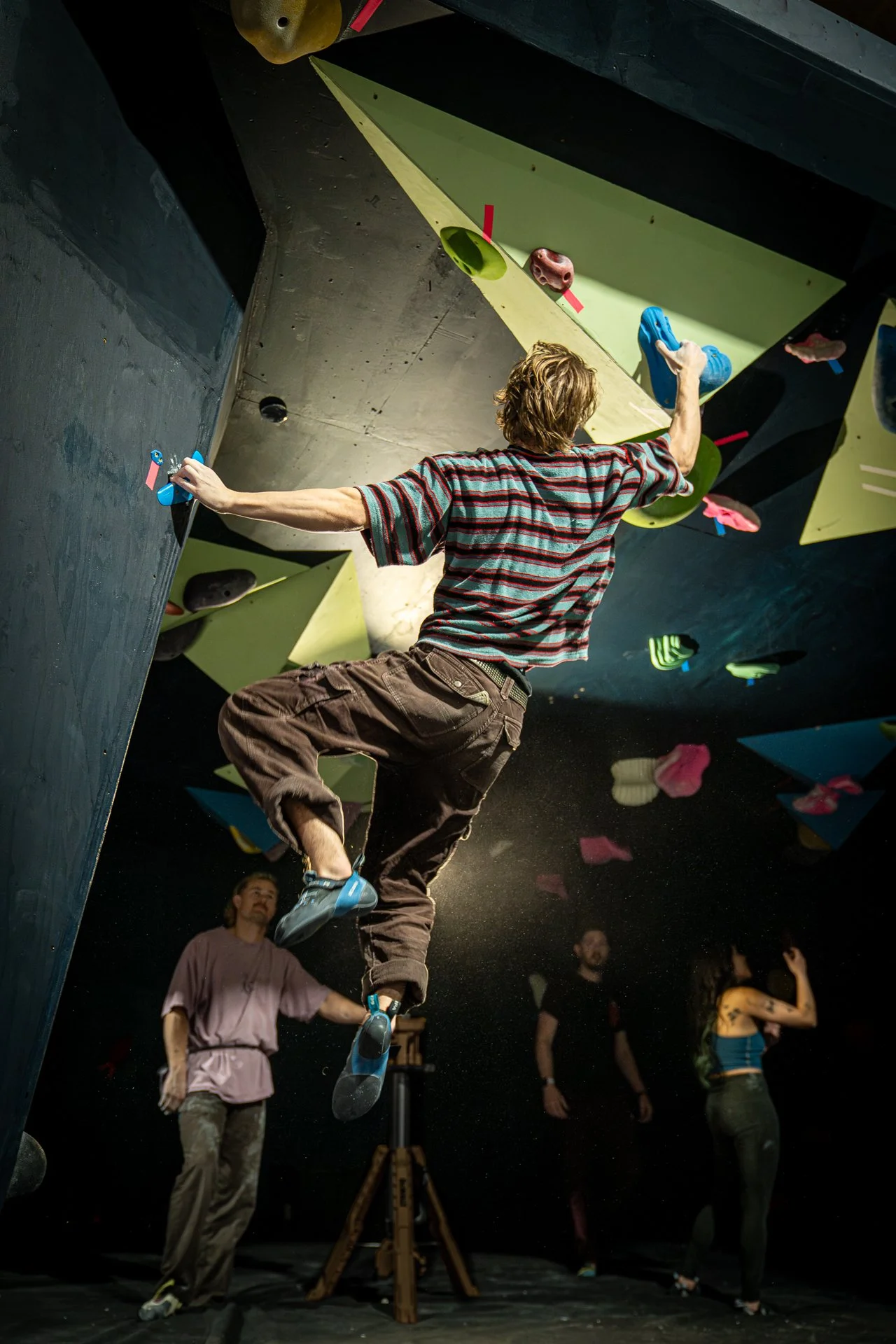 Person bouldering on an indoor climbing wall, with other climbers and spectators in the background.