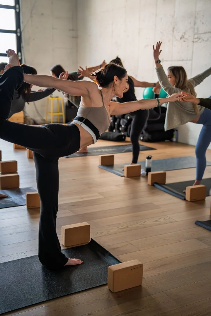 People practicing yoga in a studio, with a woman in the foreground doing a balance pose with arms outstretched, standing on one leg on a yoga mat.