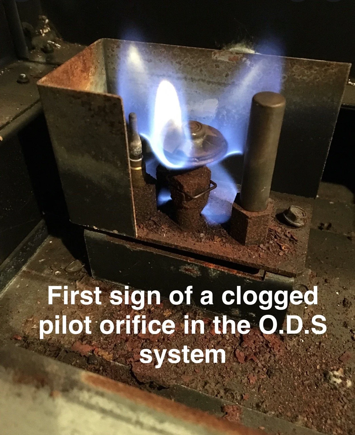 Close-up of a rusted pilot orifice with a blue flame, indicating a clogged or faulty pilot orifice in a gas system, with the text "First sign of a clogged pilot orifice in the O.D.S system" overlaid.