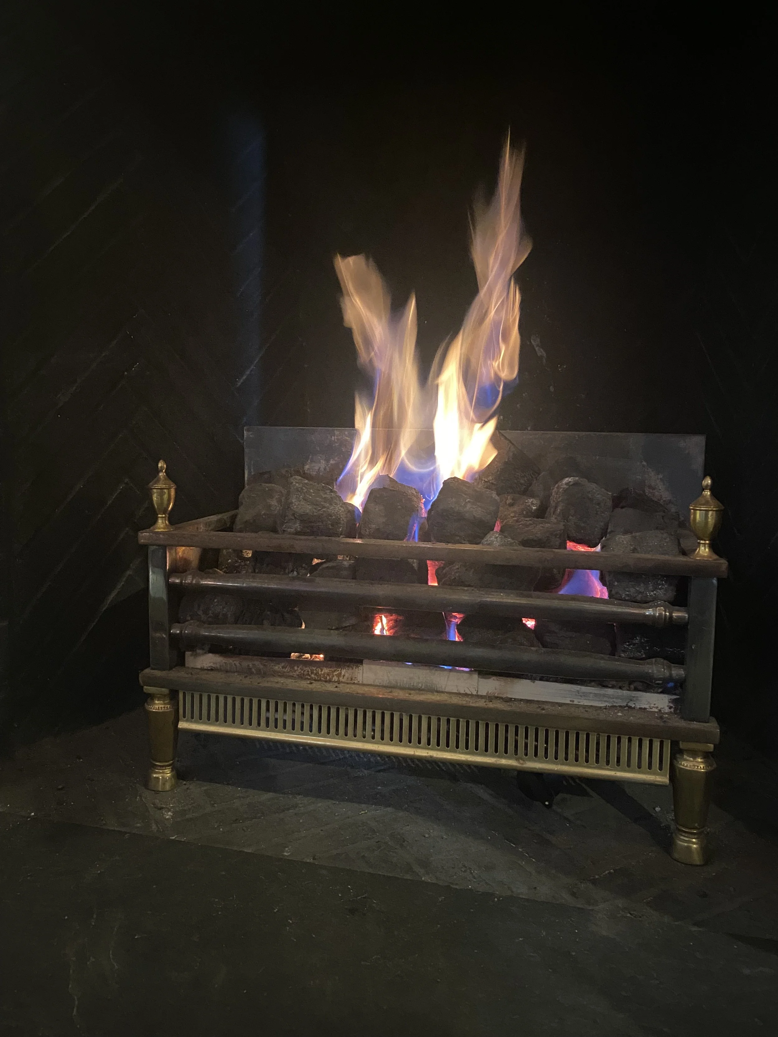 A lit gas fireplace with orange and purple flames, surrounded by black interior walls and a metal and brass frame with decorative finials.