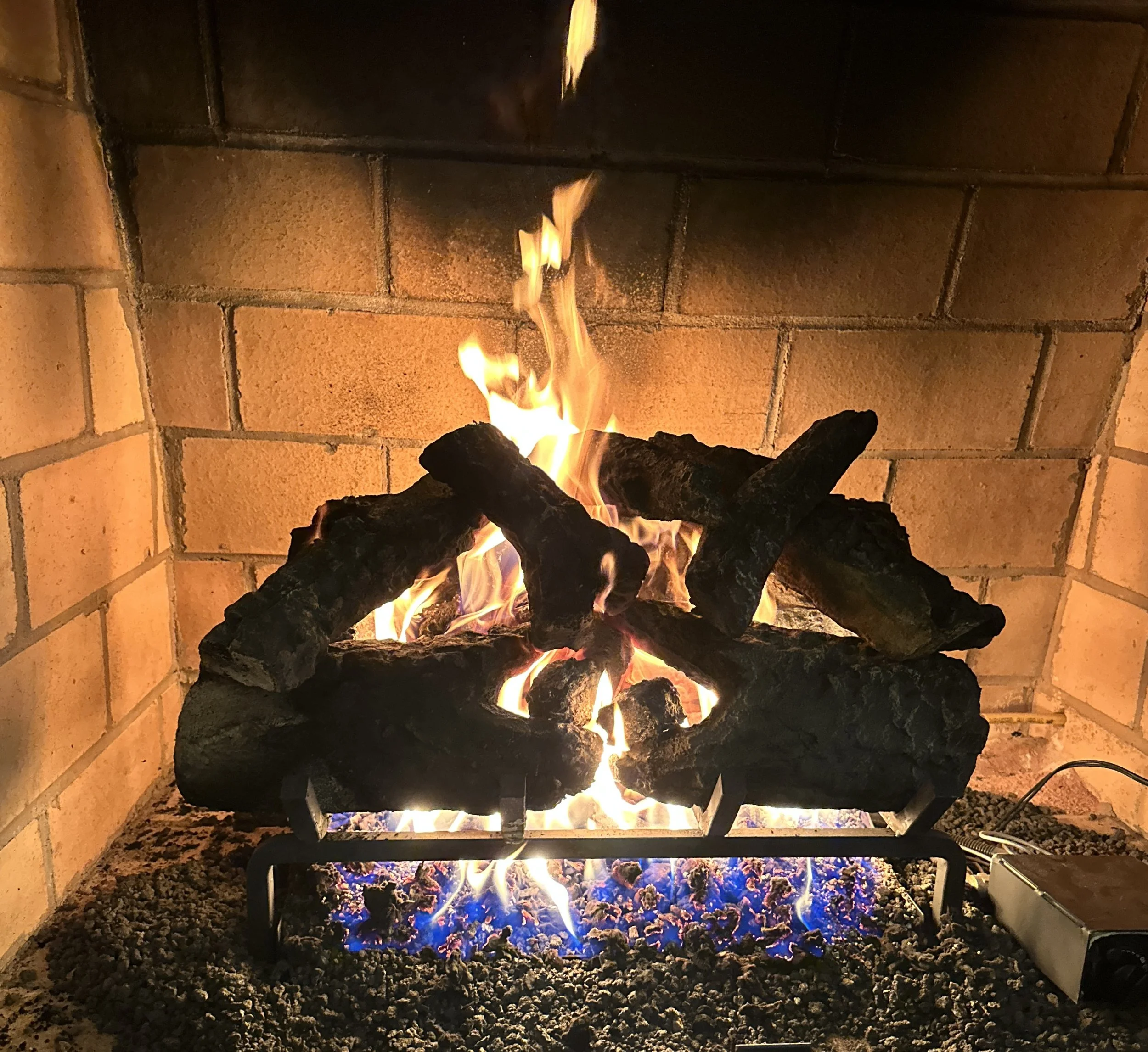 A fireplace with burning logs and bright flames, surrounded by a brick wall and grill grate at the bottom.