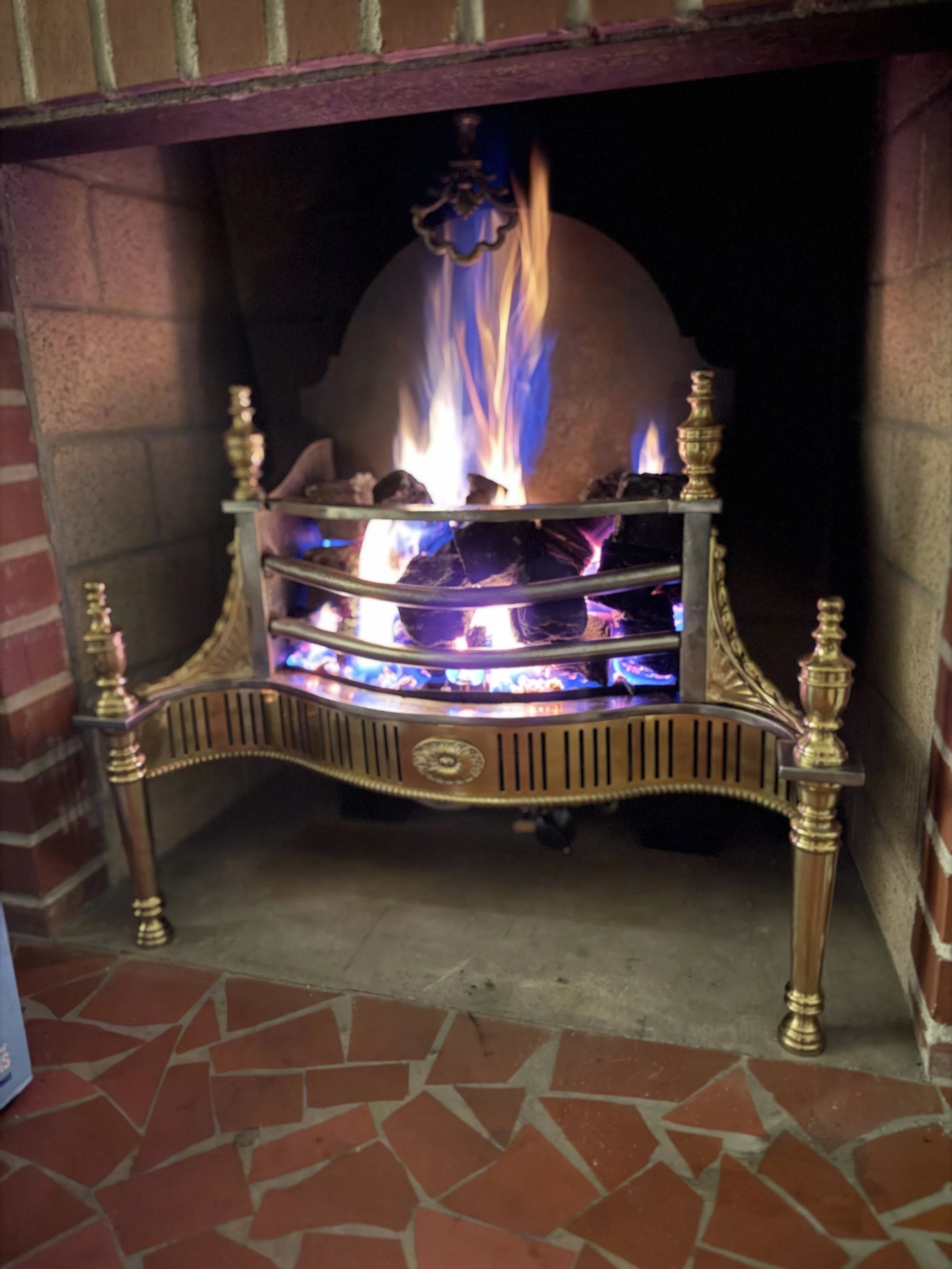 A fireplace with a metal grate and golden decorative elements, with a fire burning inside. The fireplace is set in a brick wall, with a tiled floor.
