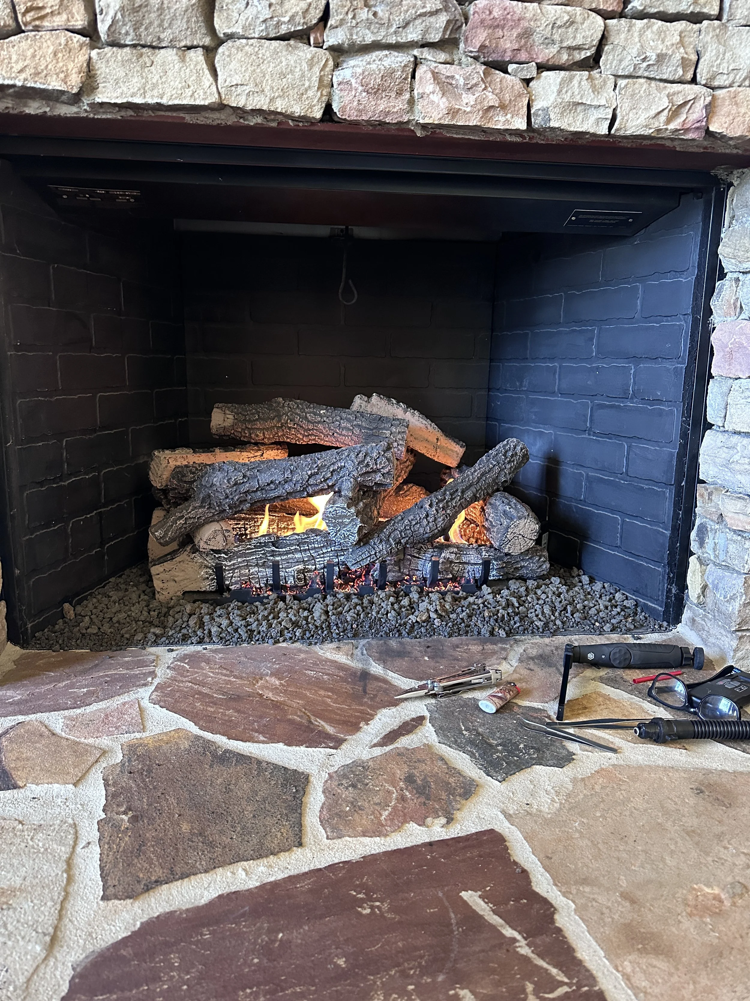 A fireplace with a stone surround and a black interior with burning logs. There are tools and a flashlight on the floor in front of the fireplace.