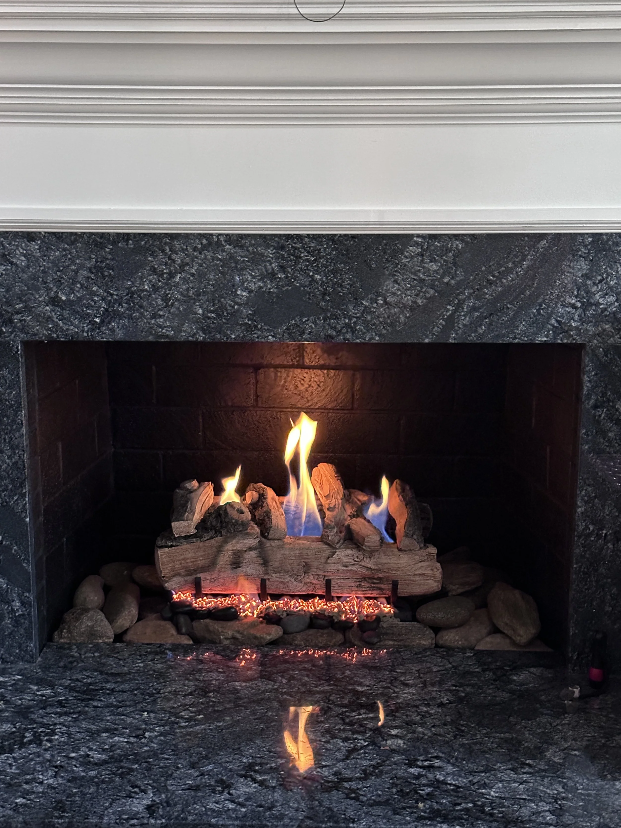 A fireplace with a brick interior and a stove top, featuring a fire with flames and logs, surrounded by a black granite mantel and decorative stones.