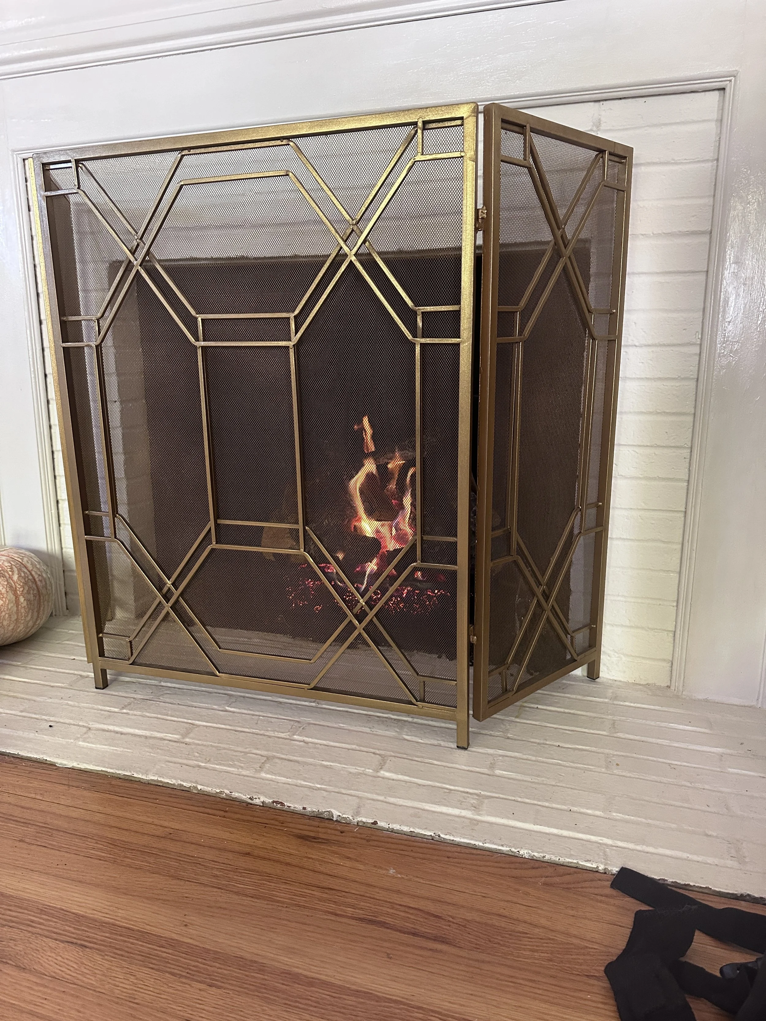 A brass-colored fireplace screen with a geometric pattern, in front of a fireplace with visible flames, placed against a white brick wall.