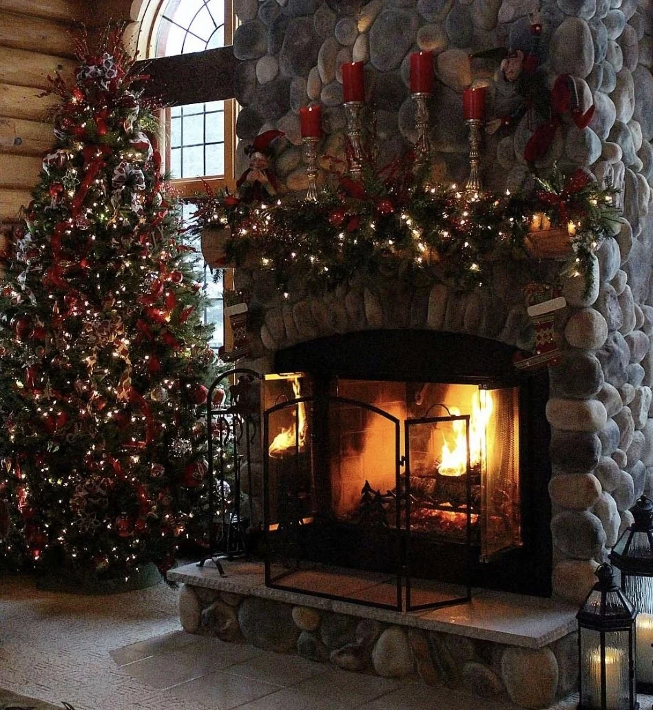 A cozy Christmas scene featuring a decorated Christmas tree with red and silver ornaments, ribbons, and lights to the left of a stone fireplace. The fireplace has a mantel decorated with ornaments, garland, and four red candles. The fire inside the f