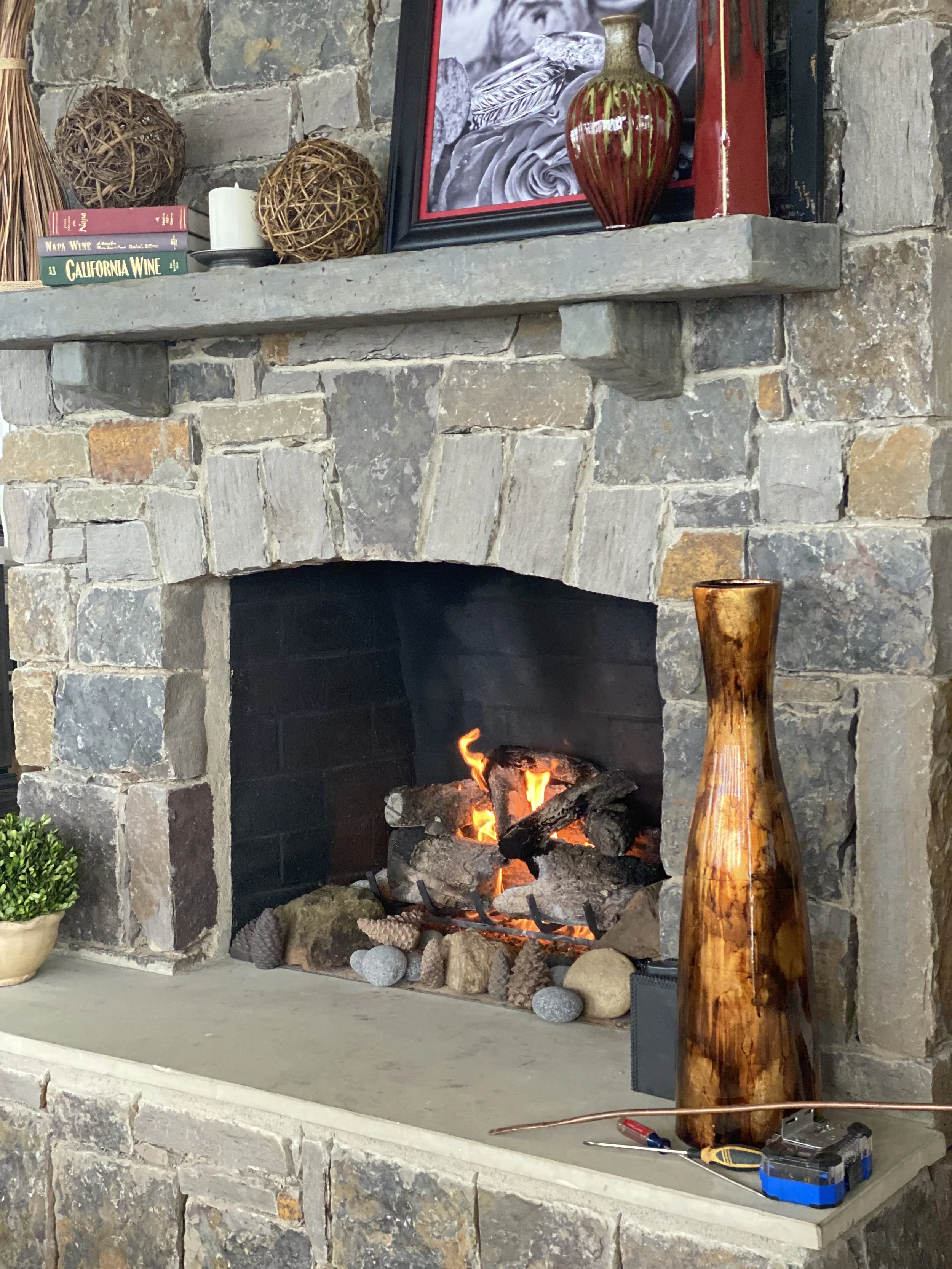 A rustic stone fireplace with a lit fire, decorative vases, stacked rocks, and a framed artwork on the mantel. There are tools and a power drill on the hearth.
