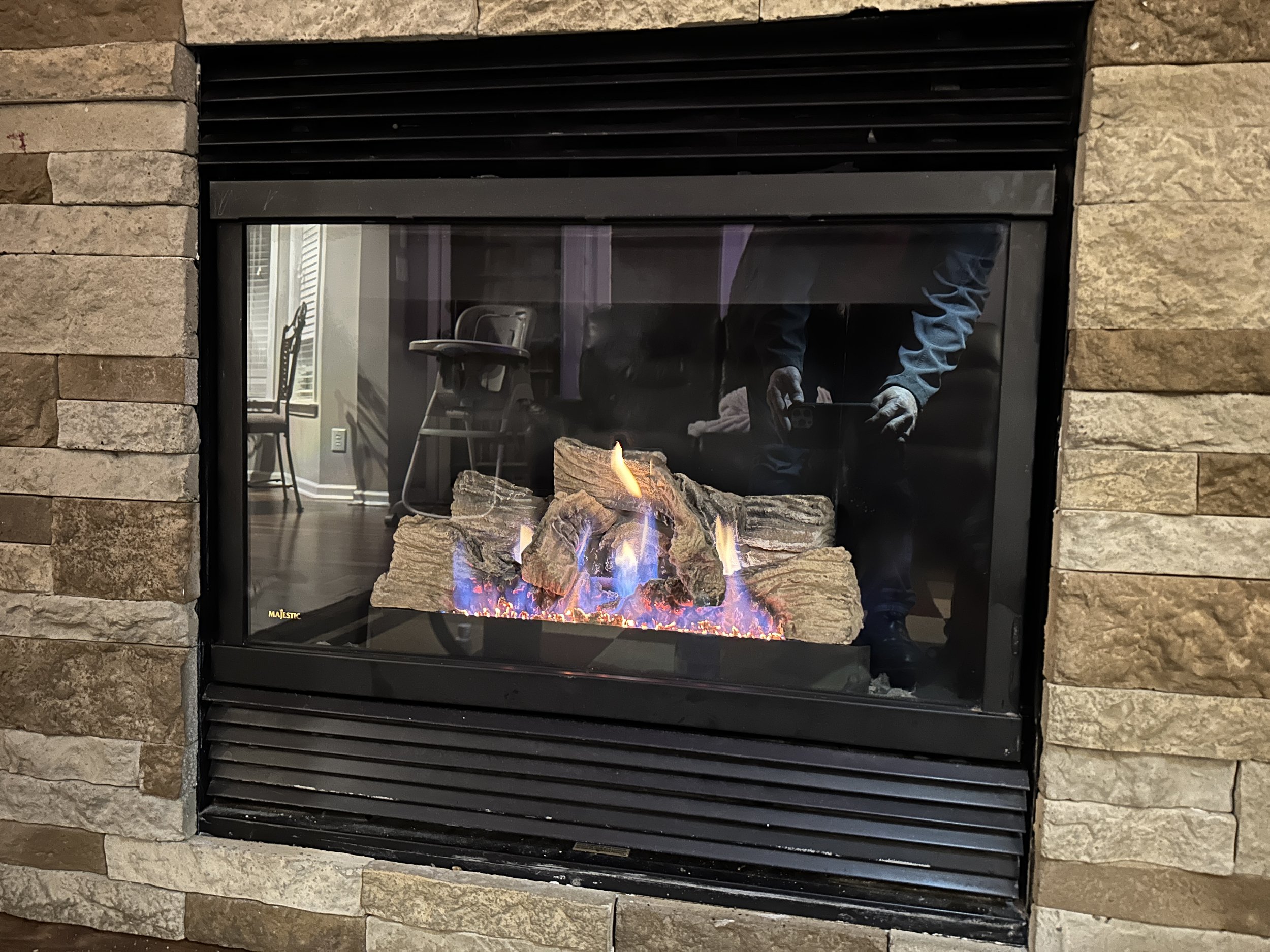 A fireplace with a stone surround, showing flames and logs inside, and a reflection of a person taking the photo.
