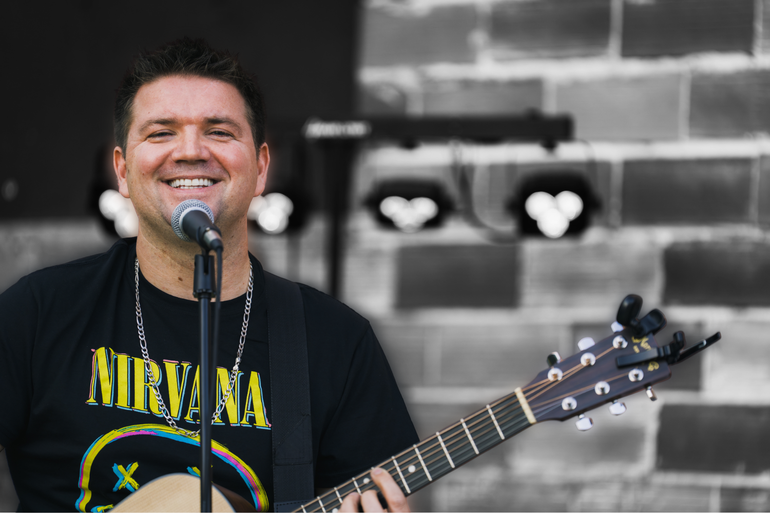 A smiling man singing into a microphone while playing an acoustic guitar in front of a brick wall.